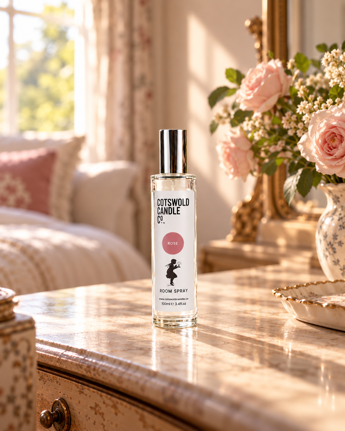Rose room spray by Cotswold Candle Co., clear glass 100ml bottle with silver cap, rose pink fragrance circle and girl logo, in a softly sunlit home setting.