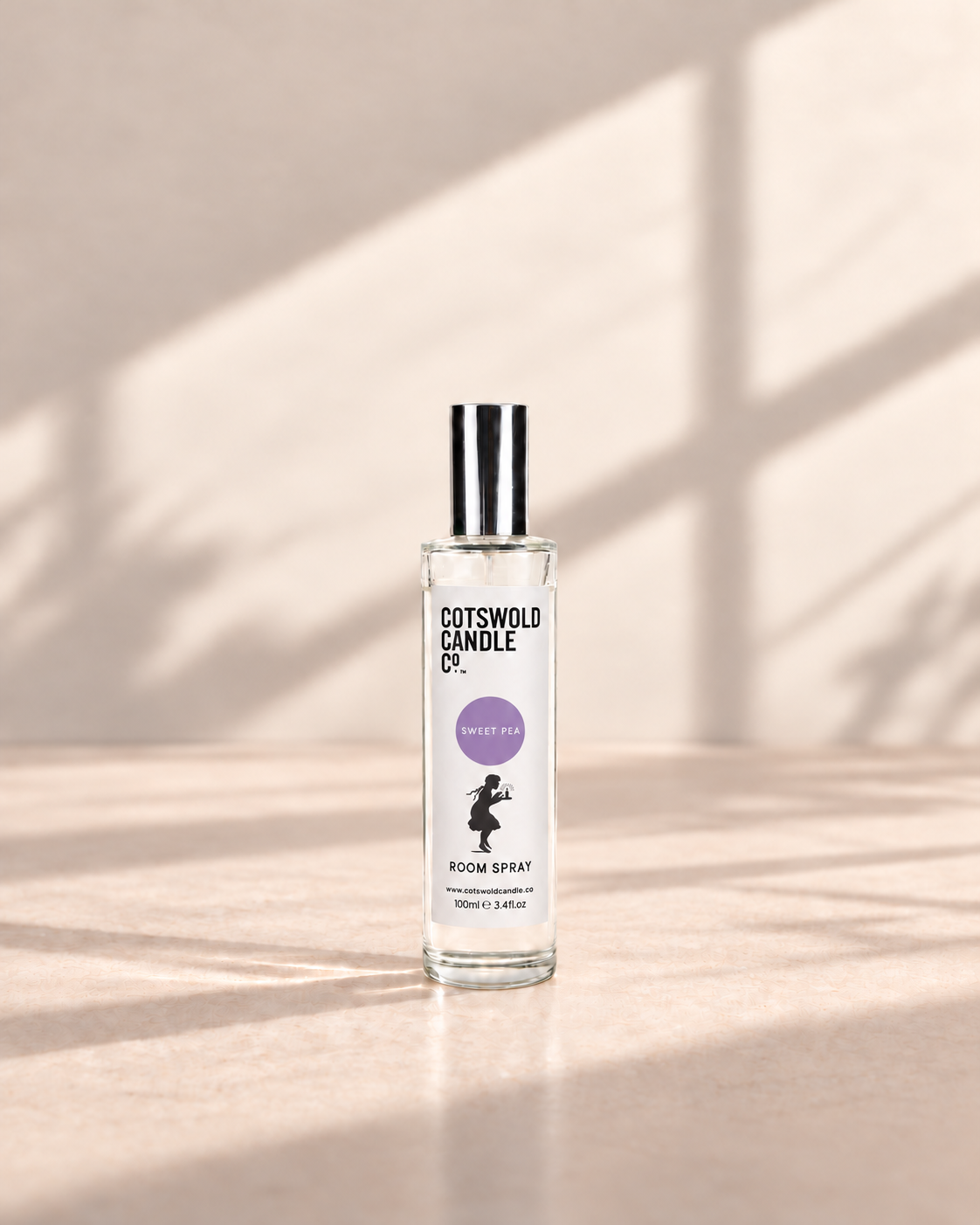 Sweet Pea room spray by Cotswold Candle Co., clear glass 100ml bottle with silver cap, lavender purple fragrance circle and girl logo, on a warm neutral studio background.