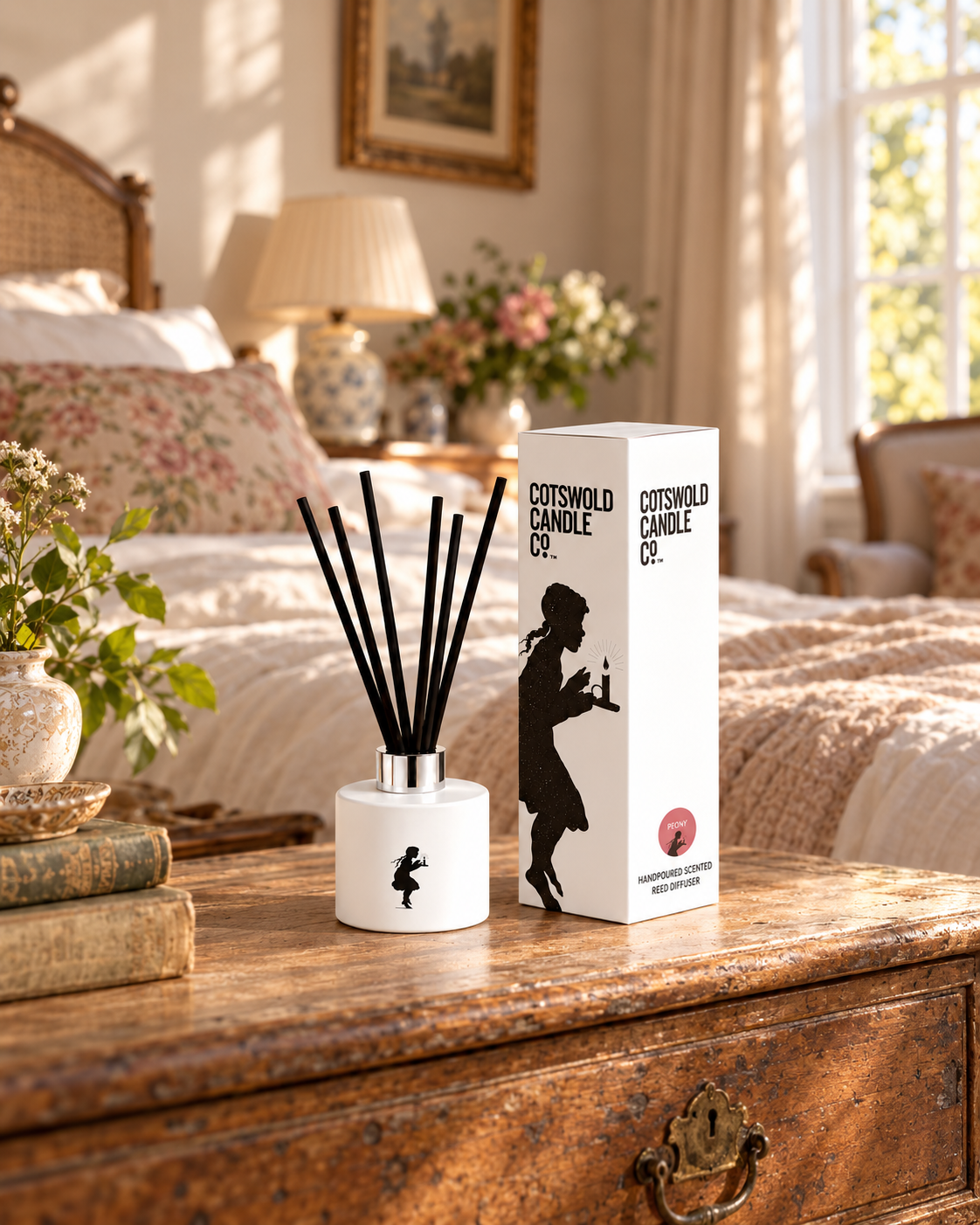 Peony reed diffuser and matching box on a wooden chest at the foot of a bed with floral cushions.
