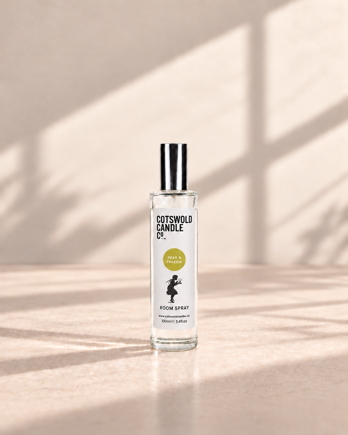 Pear & Freesia room spray by Cotswold Candle Co., clear glass 100ml bottle with silver cap, soft yellow green fragrance circle and girl logo, on a warm neutral studio background.