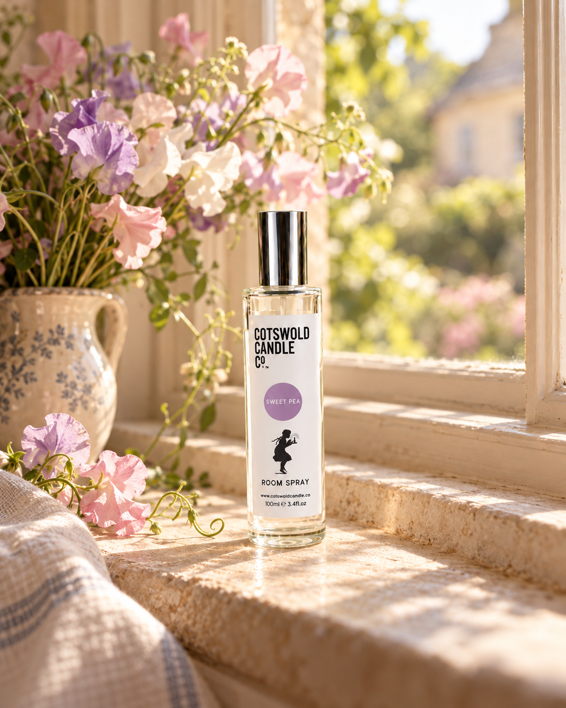 Sweet Pea room spray by Cotswold Candle Co., clear glass 100ml bottle with silver cap, lavender purple fragrance circle and girl logo, in a softly sunlit home setting.