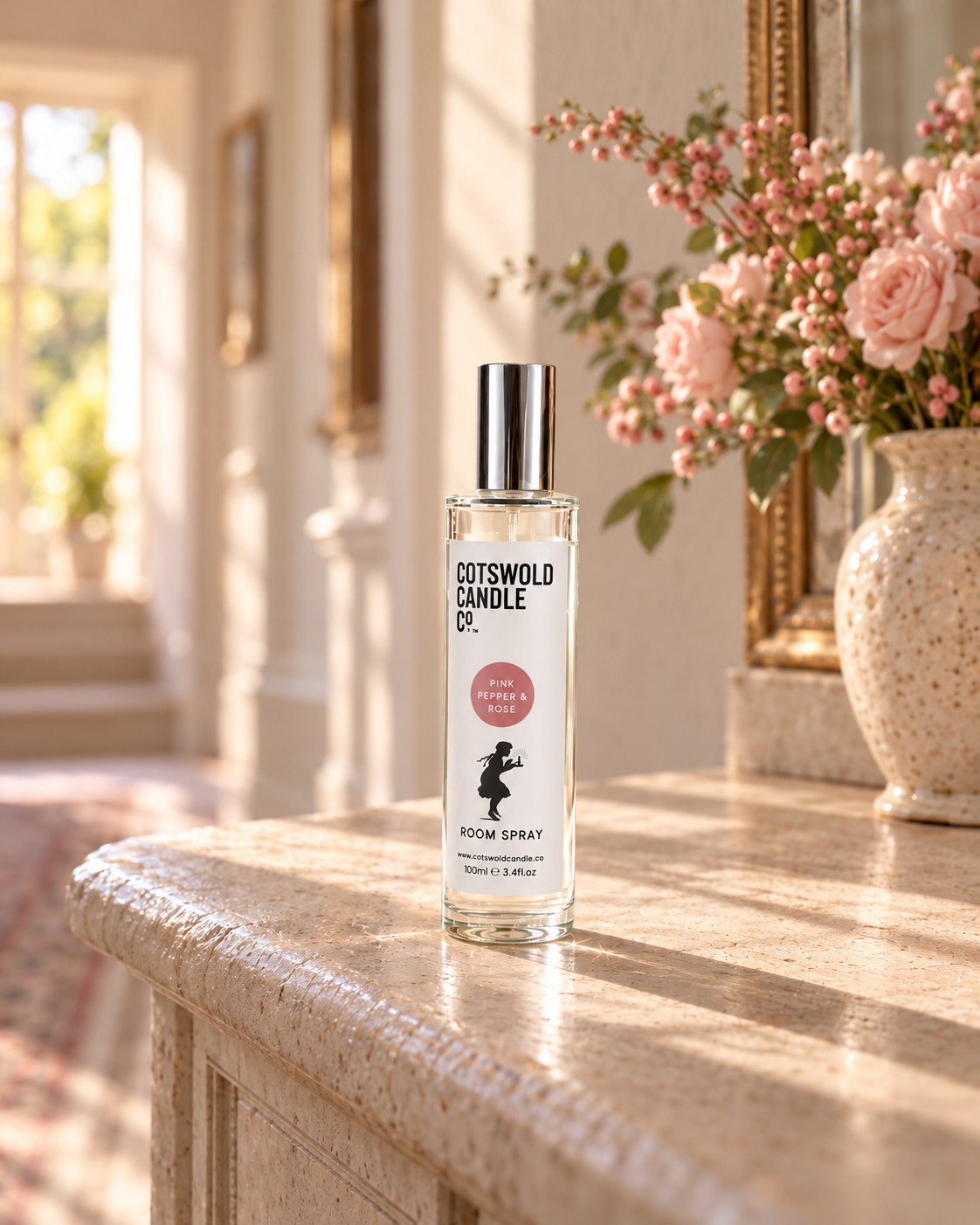 Pink Pepper & Rose room spray by Cotswold Candle Co., clear glass 100ml bottle with silver cap, dusty pink fragrance circle and girl logo, in a softly sunlit home setting.