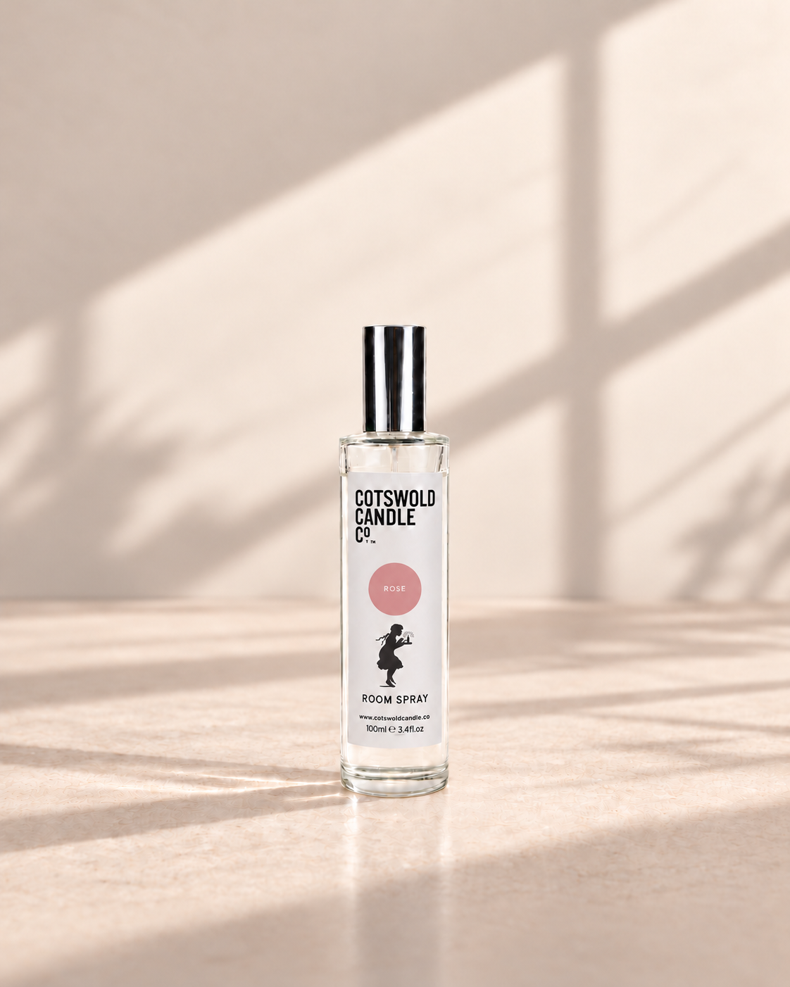 Rose room spray by Cotswold Candle Co., clear glass 100ml bottle with silver cap, rose pink fragrance circle and girl logo, on a warm neutral studio background.