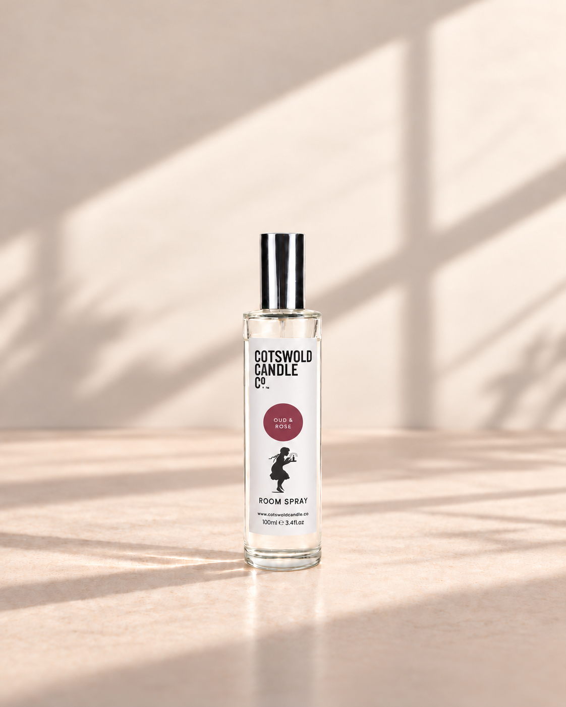 Oud & Rose room spray by Cotswold Candle Co., clear glass 100ml bottle with silver cap, burgundy fragrance circle and girl logo on a warm neutral background.