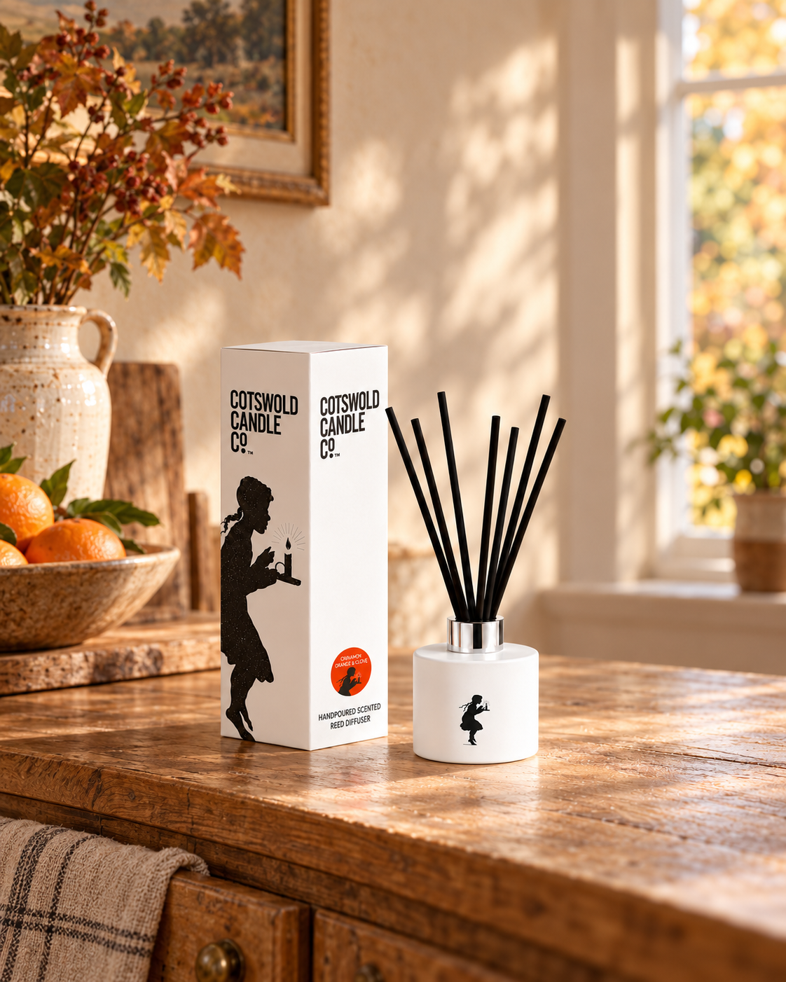 Cinnamon Orange & Clove reed diffuser and matching box on a wooden kitchen counter beside oranges and autumn foliage.