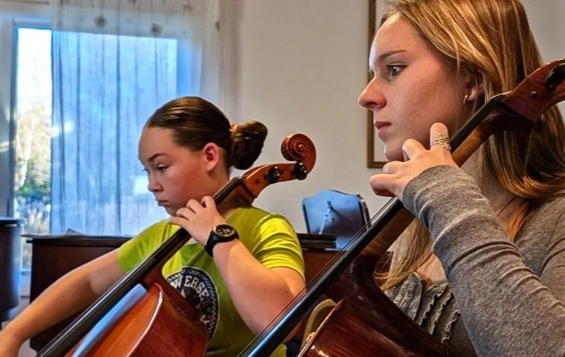 Two young women are playing cellos indoors, with a window and blue curtains in the background.