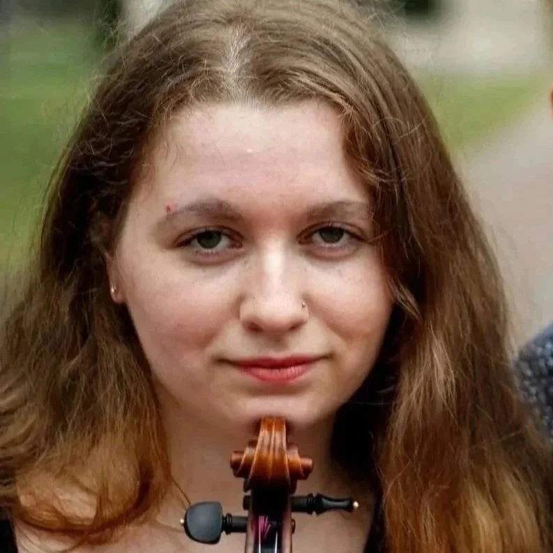 Young woman with long red hair holding a violin outdoors, next to another young person partially visible.