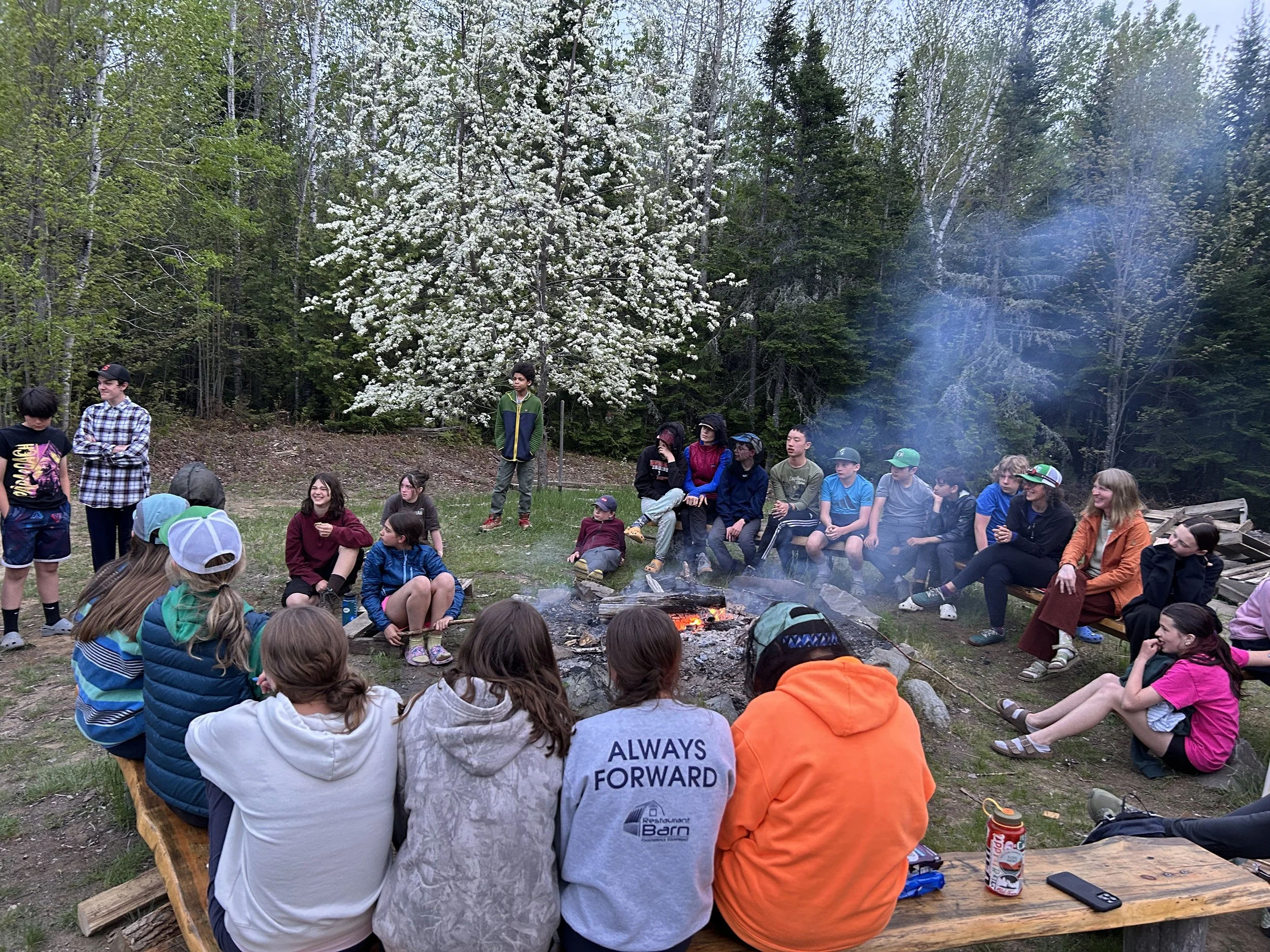 A group of middle school outdoor clubs, talking around a campfire. 