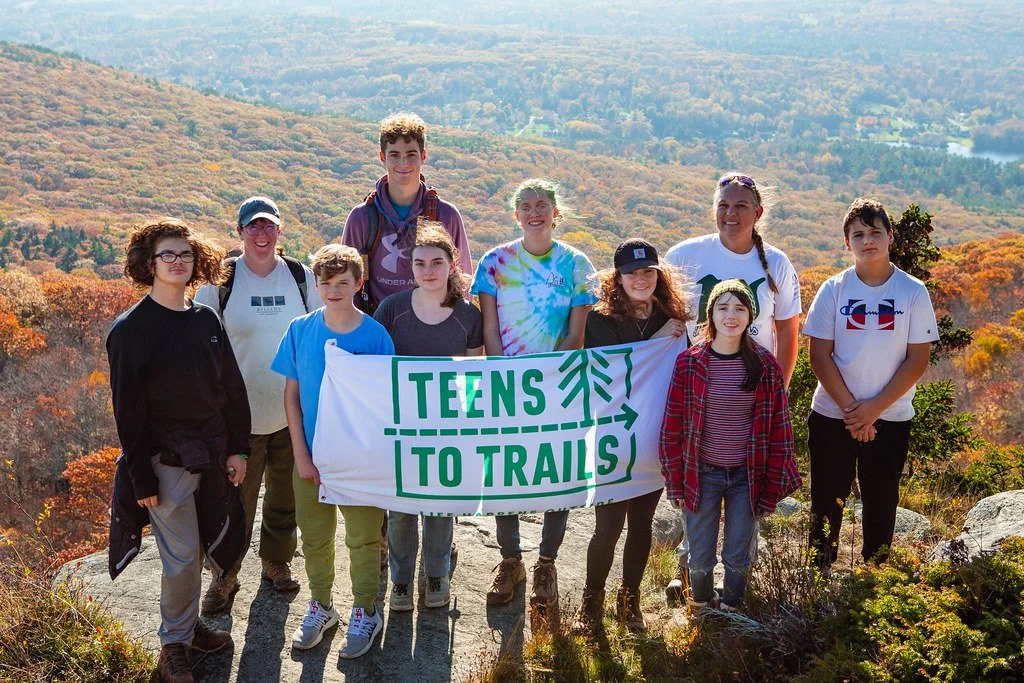 Group of young people holding a banner that reads 'Teens to Trails' during a hike on a mountain trail with scenic fall foliage in the background.