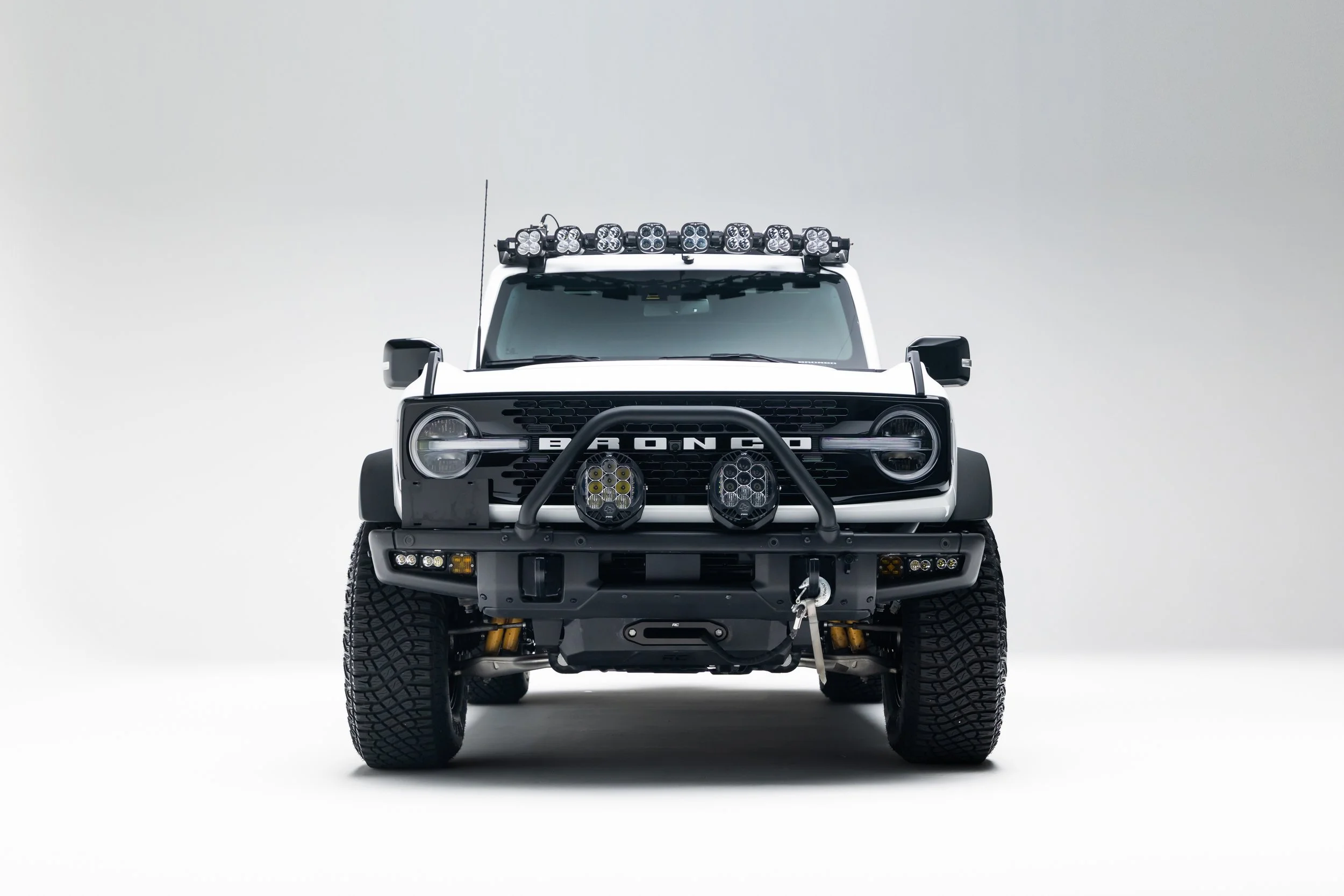 A white Ford Bronco SUV is photographed head-on in a plain studio setting. It has a black front bumper with a winch hook, auxiliary lights on the bumper and roof rack, wide off-road tires, and visible BRONCO grille lettering.