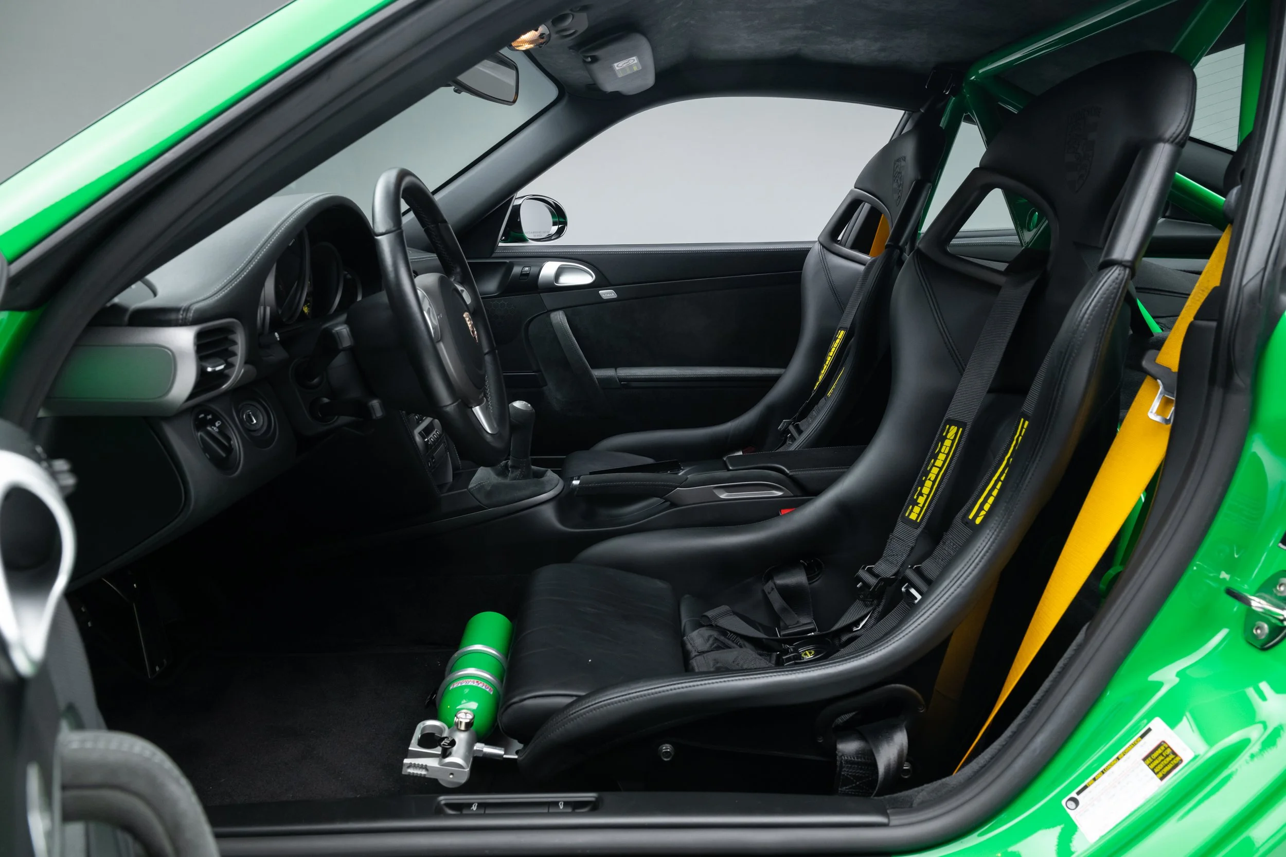 Interior of a 2008 Porsche 911. Features two racing seats with harnesses, a central console, and a steering wheel. A fire extinguisher is positioned near the driver's seat. Dashboard displays are visible.