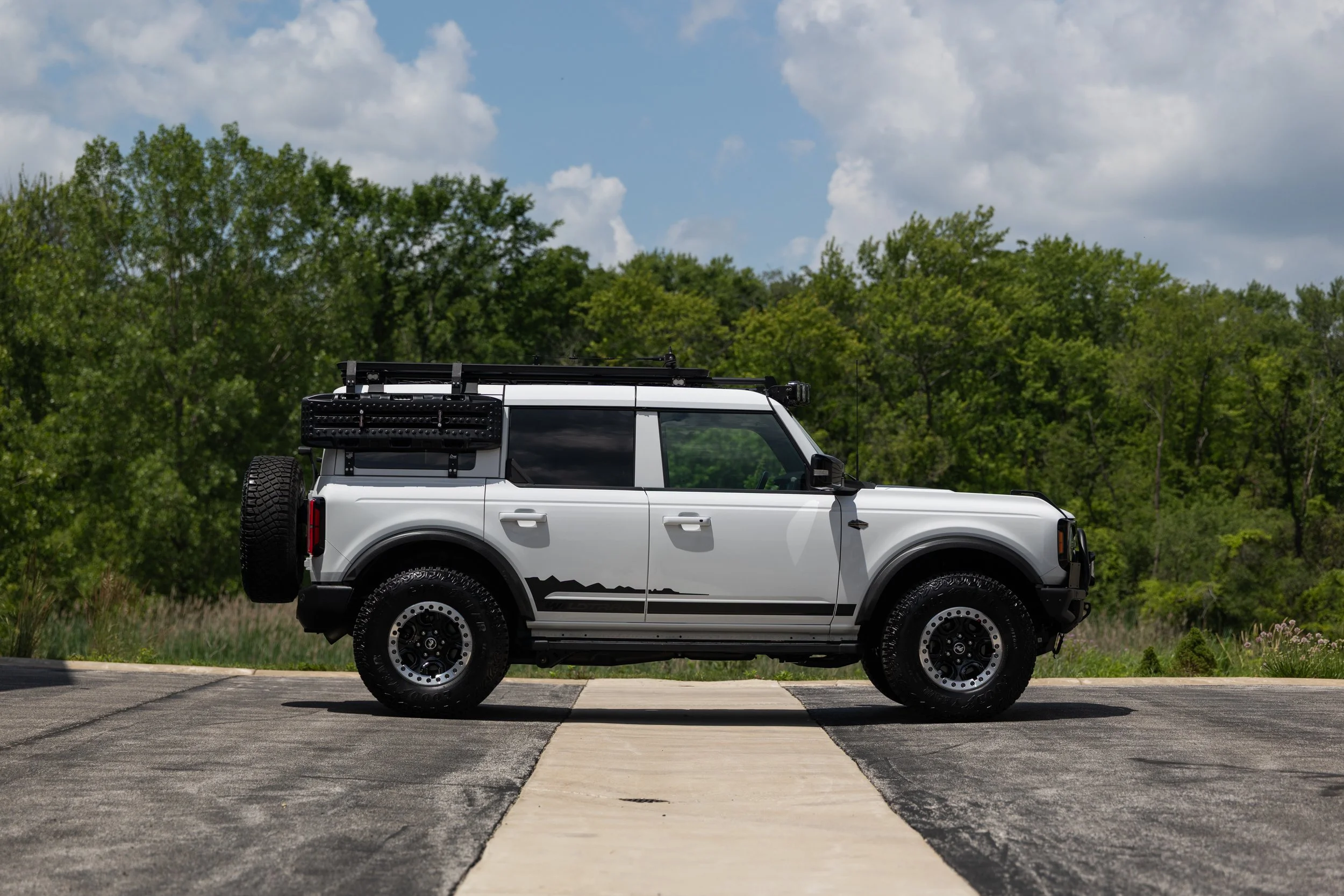 A white four-door Ford Bronco is shown in full side profile on a paved area with trees and cloudy sky in the background. It has black fender flares, off-road tires, a rear-mounted spare tire, roof rack, and side-mounted overland gear.