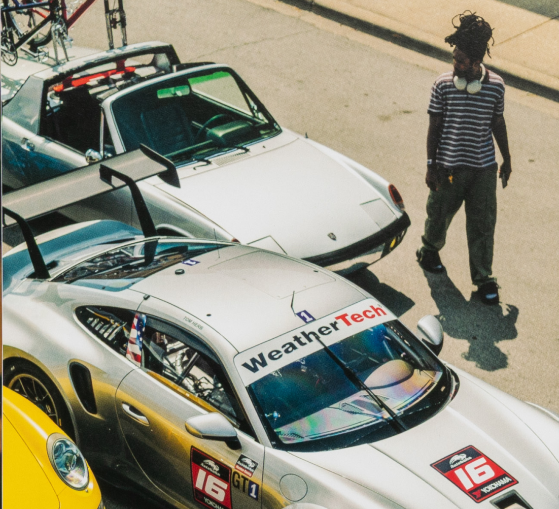 Overhead exterior view of parked cars at an outdoor event; silver race car with large rear wing and branding, with a person standing nearby.