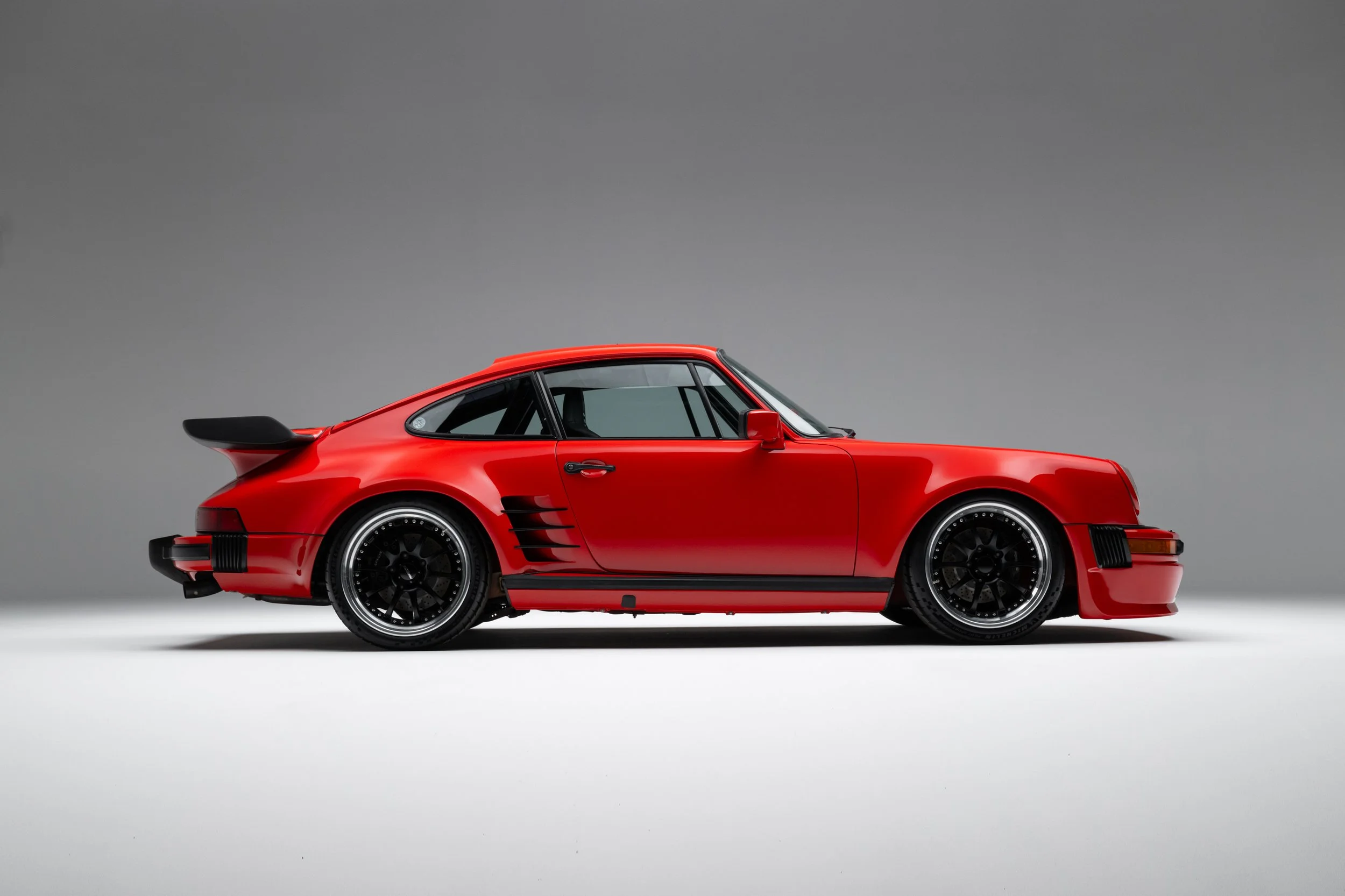 Porsche 911 (1985) featuring a red exterior, distinct rear wing, black alloy wheels, and racing stripes on the side. The vehicle showcases a streamlined profile and classic design elements typical of the 911 model.