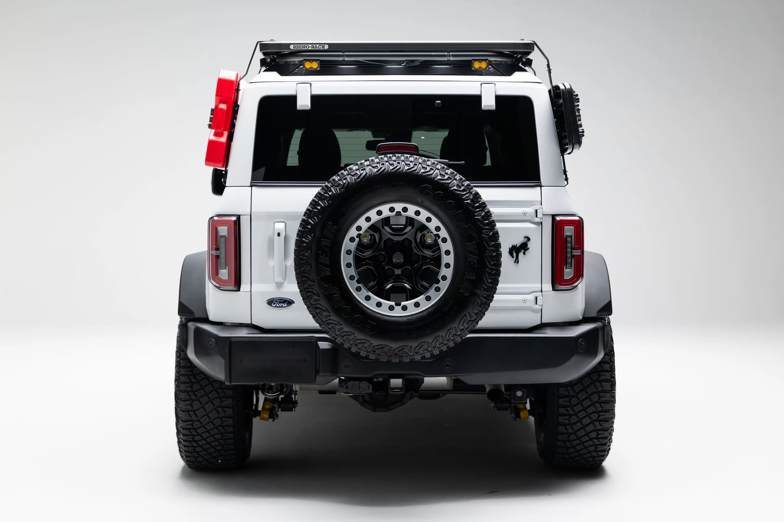 A white Ford Bronco four-door SUV is shown from the rear in a studio setting on a plain light background. The rear-mounted spare tire, roof rack with lighting, side-mounted gear, black bumper, and vertical taillights are visible.
