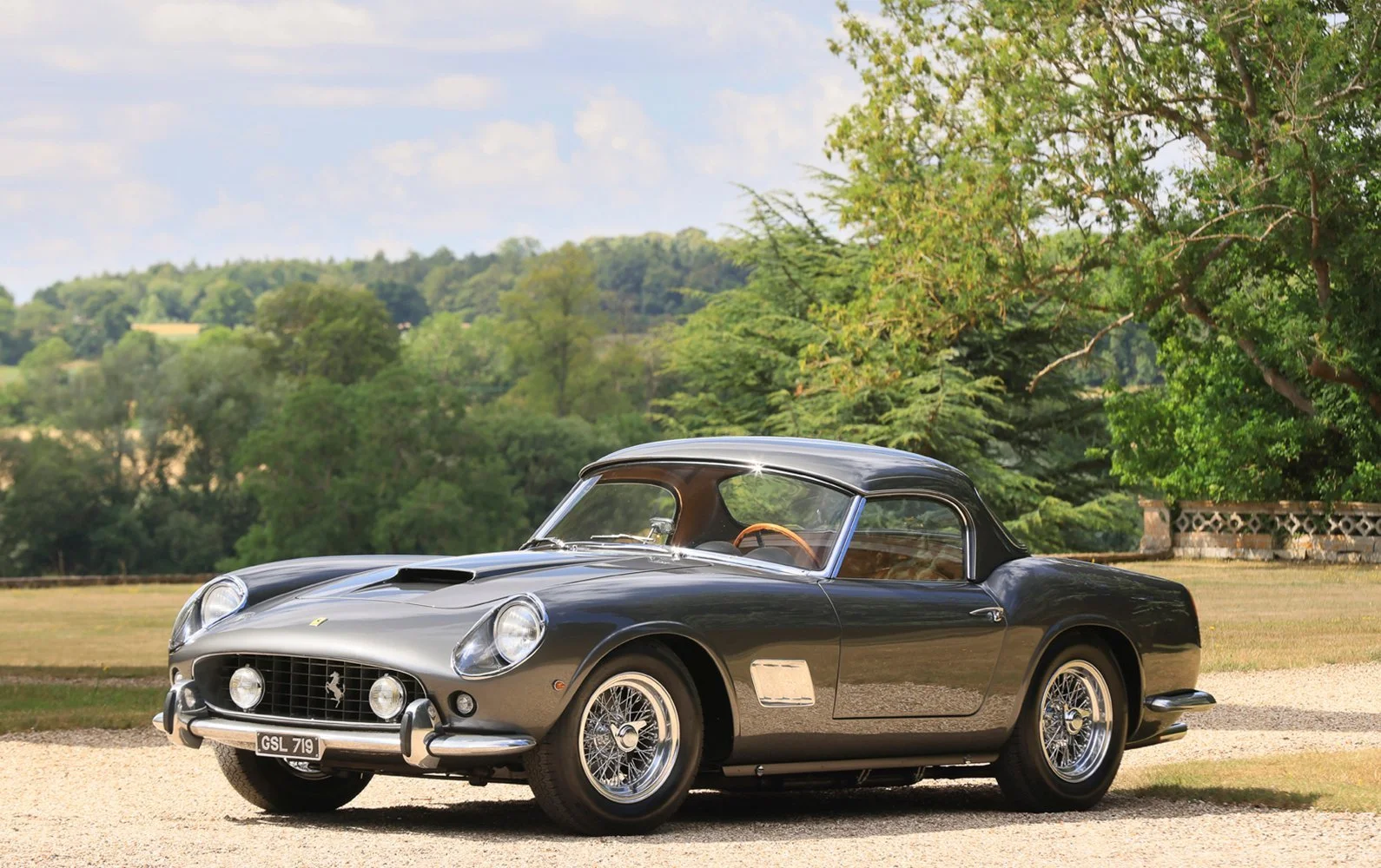 Grey classic Ferrari with a hardtop parked on a gravel drive.