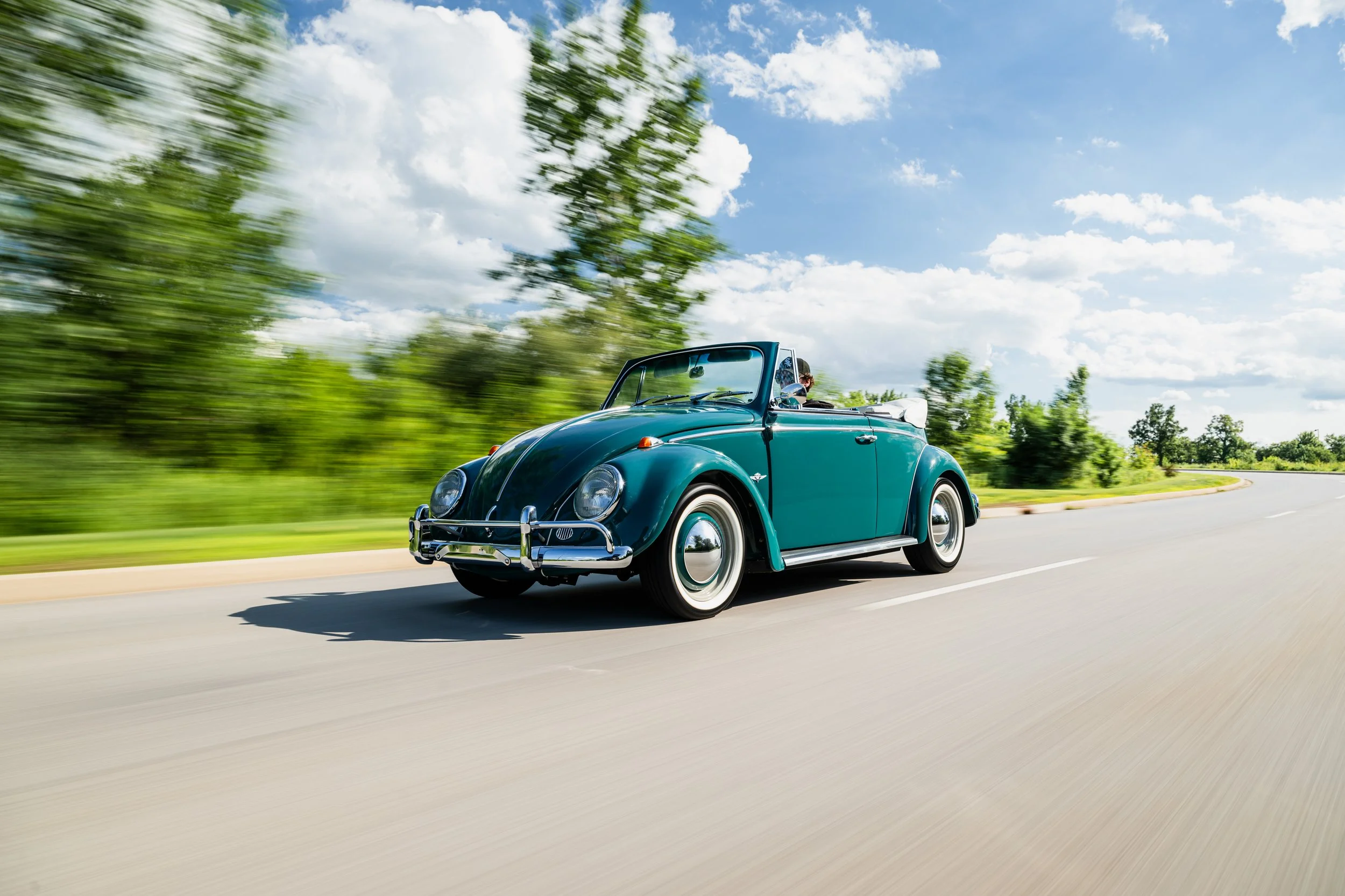 A teal 1964 Volkswagen Beetle convertible is driving on a paved road with blurred green trees and a partly cloudy sky in the background. The car is shown from a front three-quarter exterior view with chrome bumpers, roun