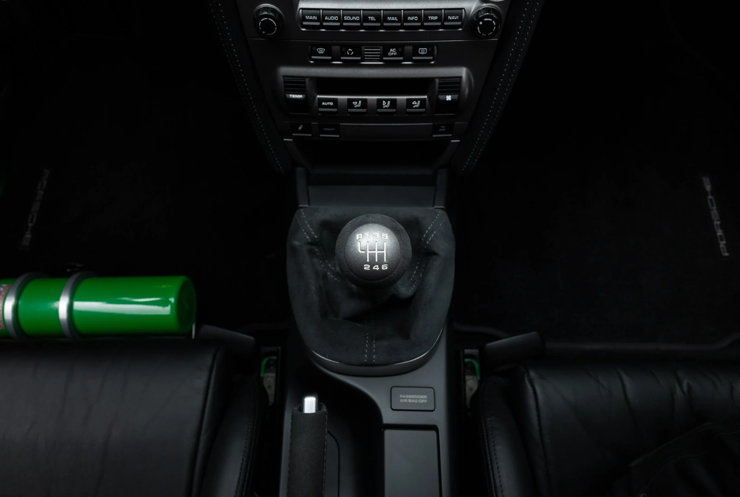 Interior view of a 2008 Porsche 911 showcasing a manual gear shift with 6-speed pattern. Black leather seats and dashboard controls are visible. A green cylinder is seen to the left of the gear shift.