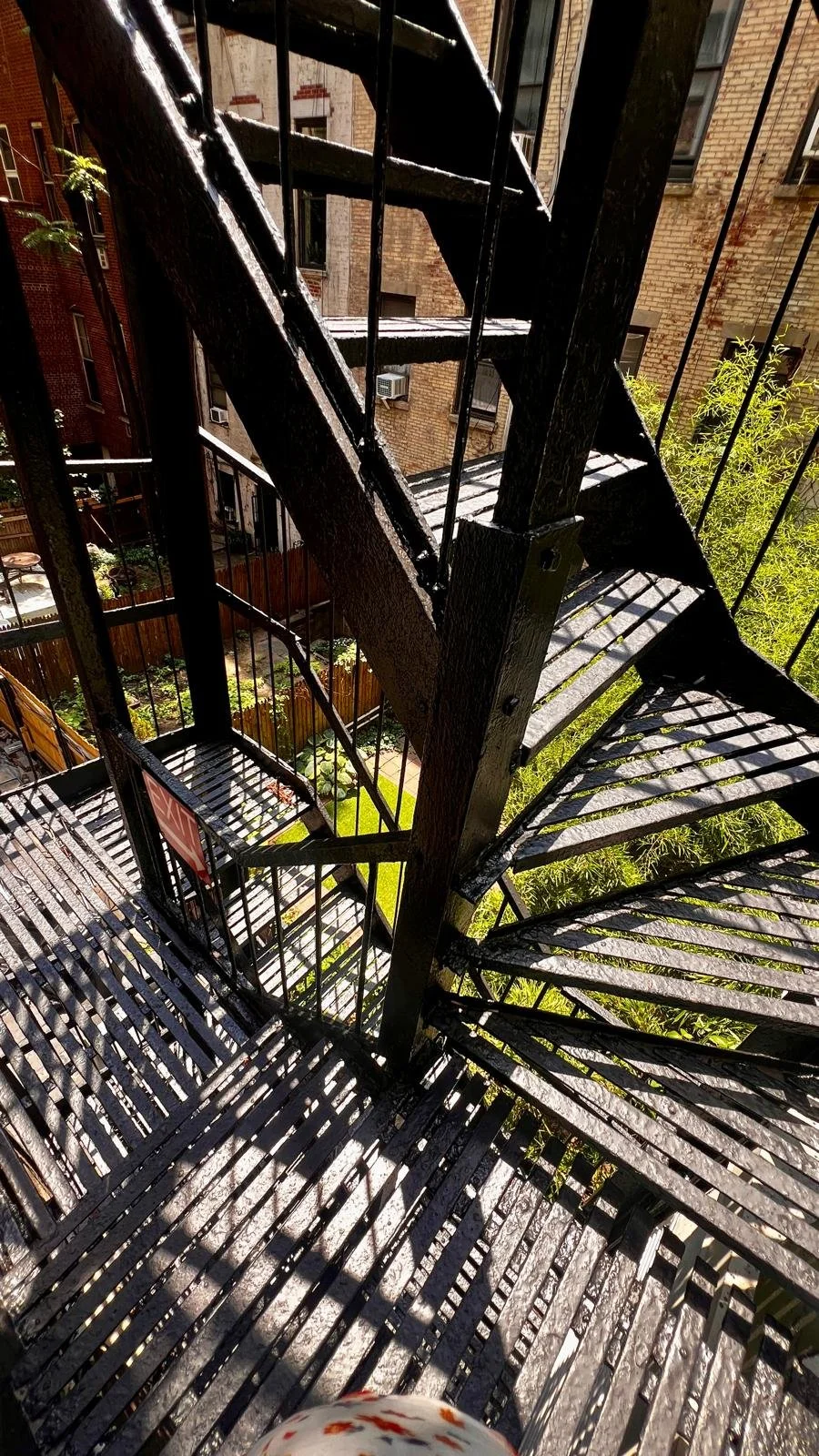 View of a black metal spiral fire escape staircase on the side of a brick building with shadows cast from the metal grates, looking down from a higher floor.