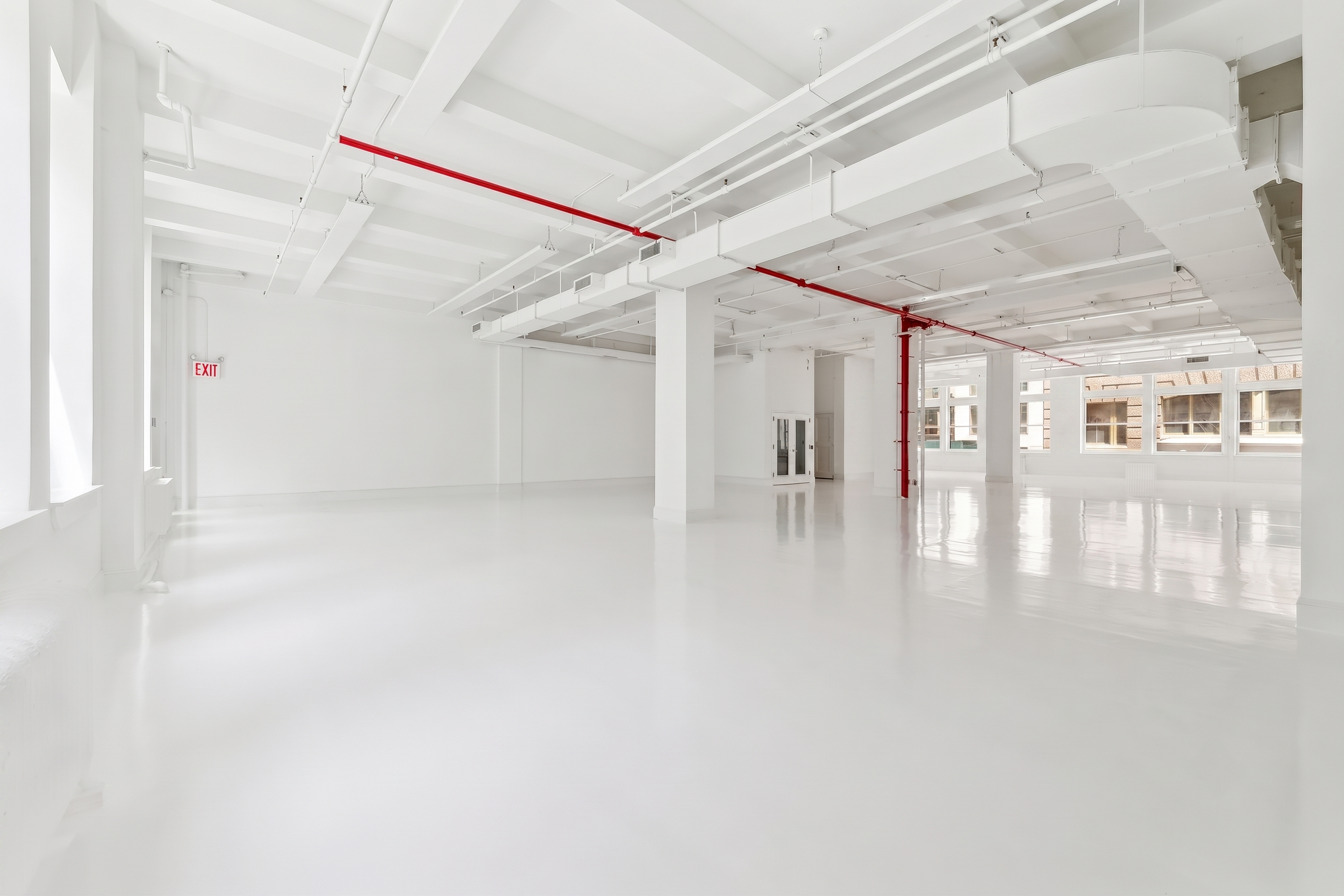 Empty, spacious room with white walls, white ceiling, and large windows letting in natural light. Exposed pipes and red sprinkler pipes run across the ceiling, and there's an exit sign on the wall. The room has a glossy white floor and appears to be in a building under renovation or construction.