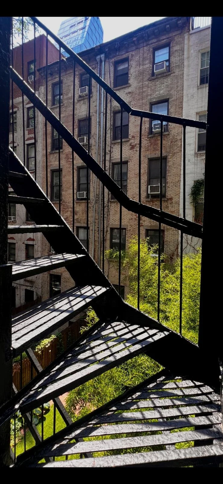View from an outdoor fire escape on a city apartment building, showing brick walls, windows with air conditioning units, and greenery below.