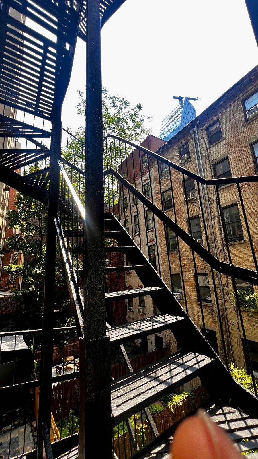View from underneath a black fire escape ladder on an urban building, with sunlight casting shadows on the stairs, neighboring brick buildings, trees, and a modern skyscraper in the background.
