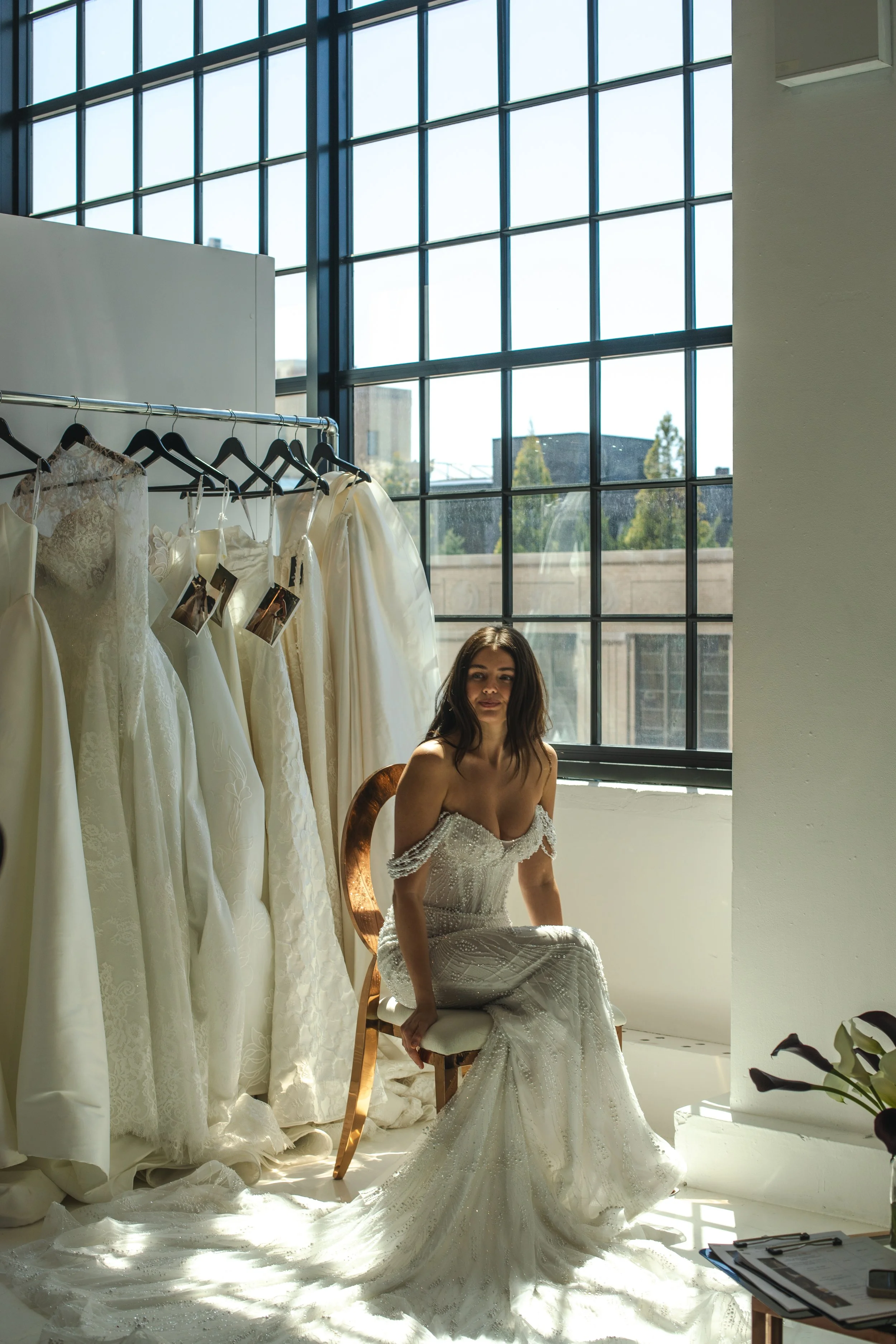 Bride in a beaded off-the-shoulder wedding gown seated by a clothing rack at Daylight Studio's NYC bridal showroom venue.