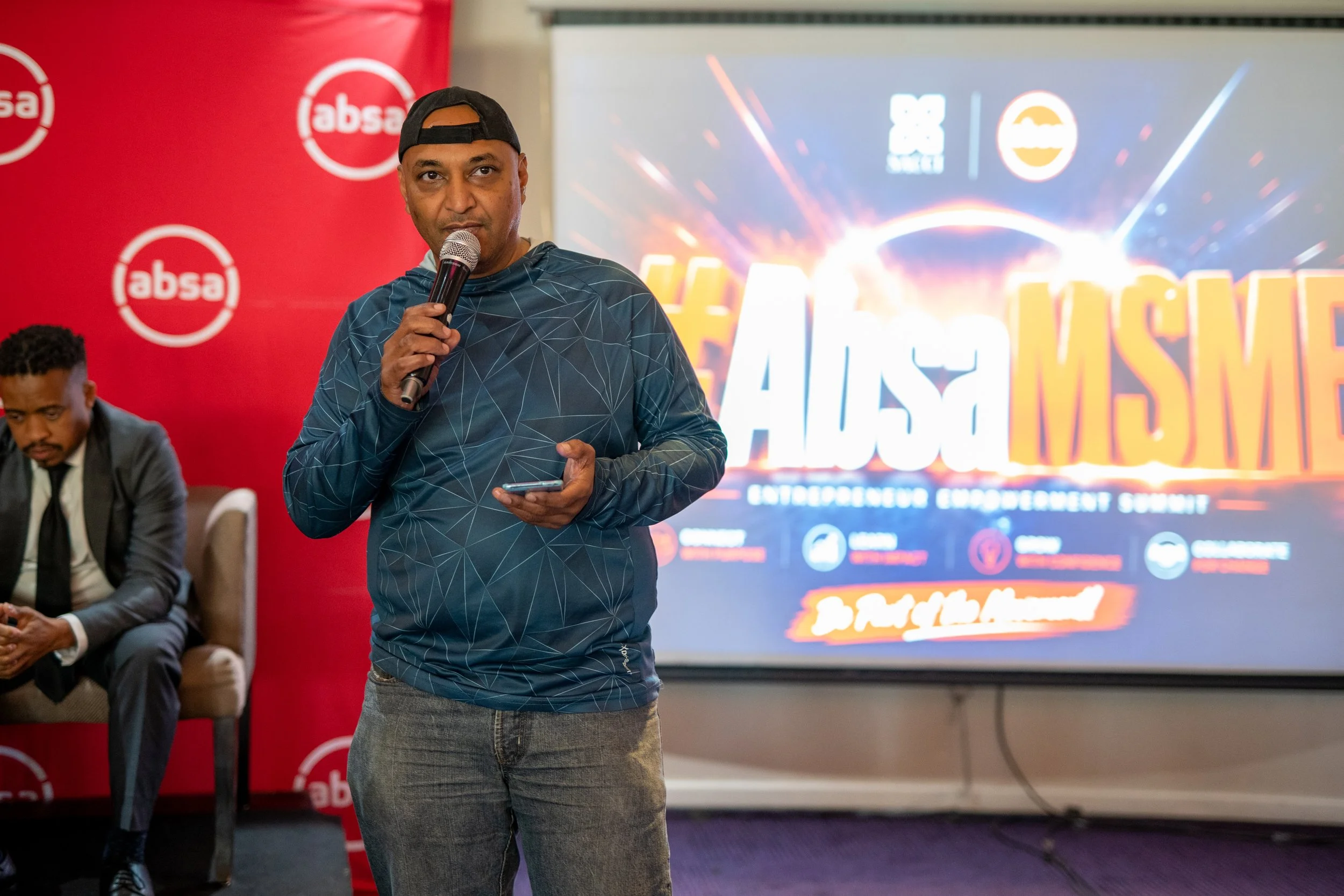 A man wearing a black cap backward, with a blue patterned hoodie and gray jeans, is holding a microphone and a phone while speaking at an event. Behind him is a large screen displaying a colorful logo, and to his left is a red backdrop with the 'absa