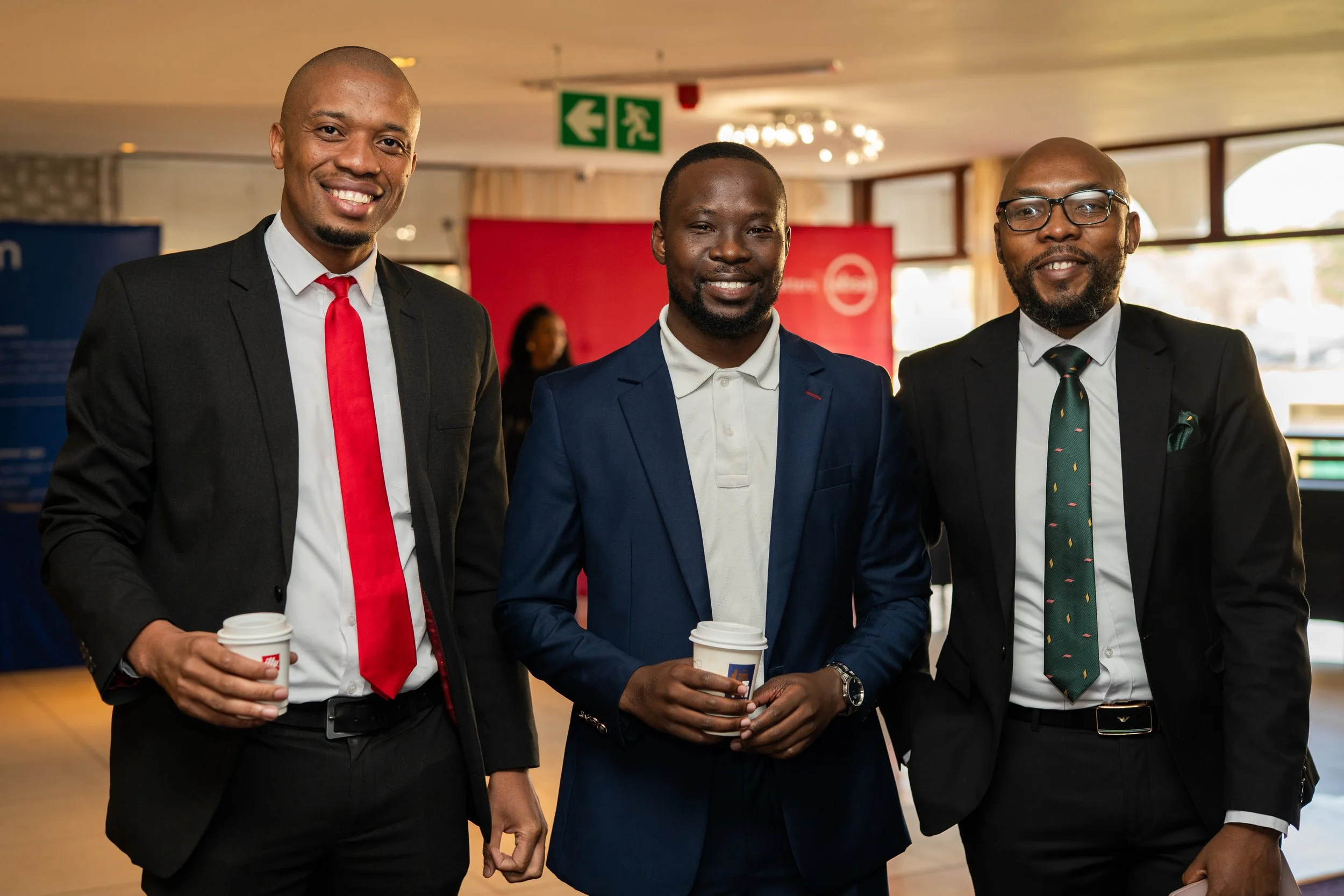 Three men dressed in business attire holding coffee cups at indoor event, smiling at camera.