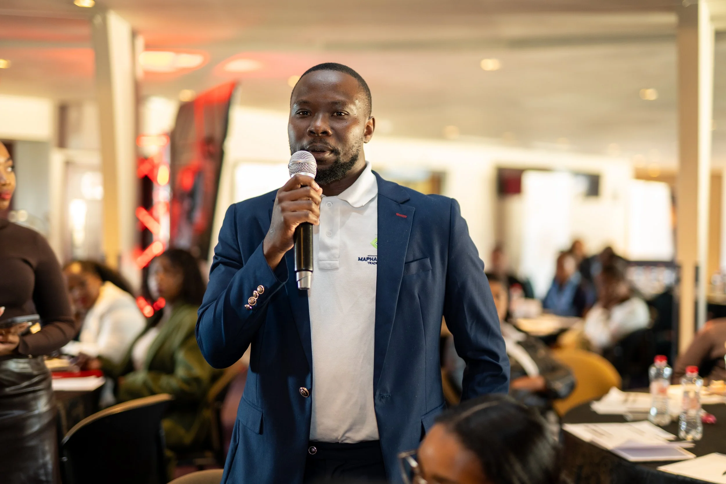 A man in a navy suit and white shirt holding a microphone speaking to an audience at an indoor event.