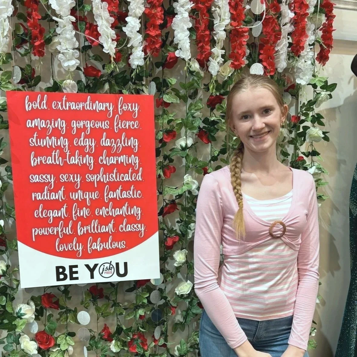 A young woman with long blonde hair styled in a braid, wearing a pink and white striped top with a pink cardigan, standing in front of a floral wall with white, red, and green flowers. There is a red sign with white cursive text next to her listing positive adjectives, and bold black text at the bottom reading "BE YOU."