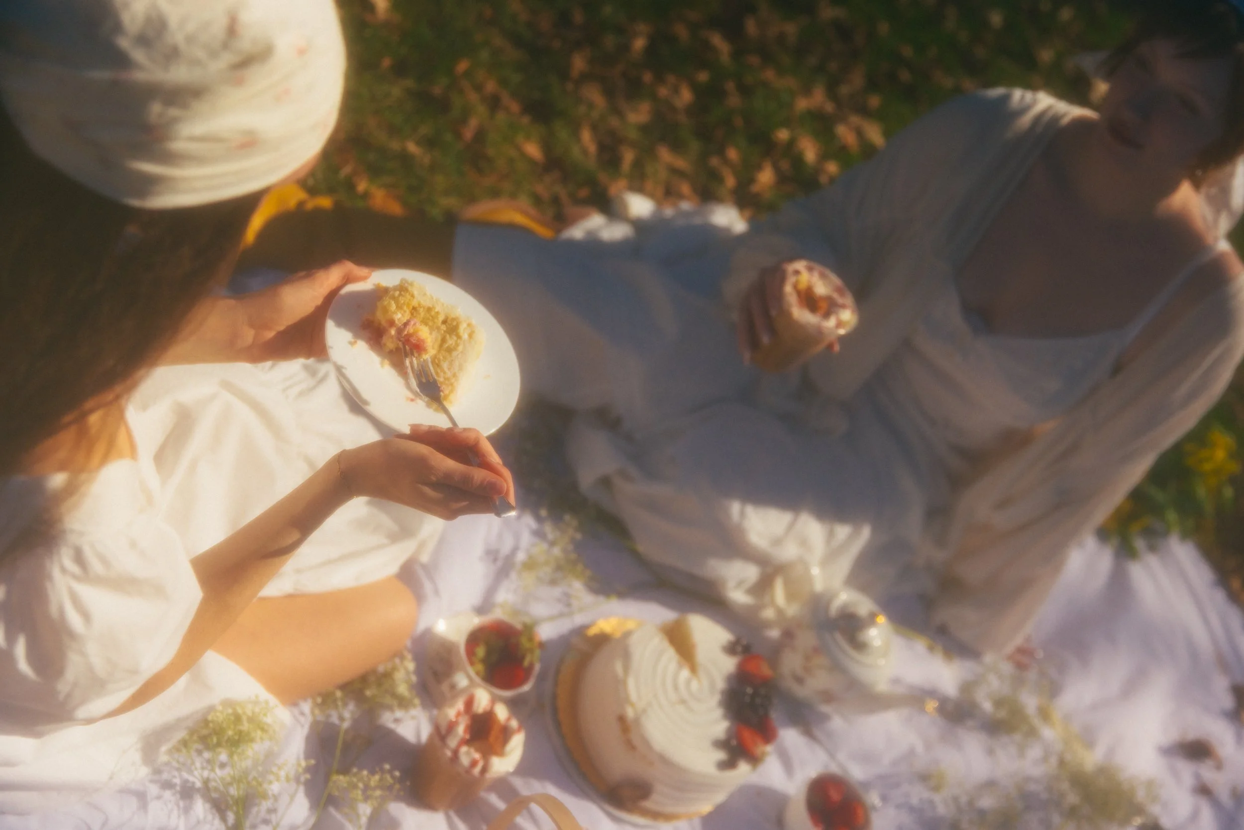 A person holding a slice of cake at an outdoor dessert table with various cakes and strawberries visible.