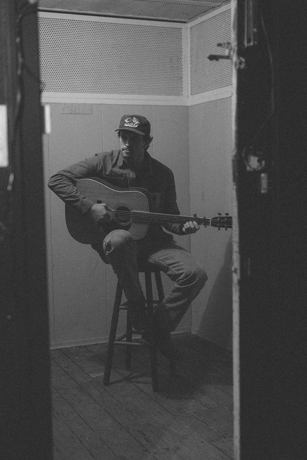 A man wearing a cap and dark clothing is sitting on a stool and playing an acoustic guitar in a small, dimly lit room.