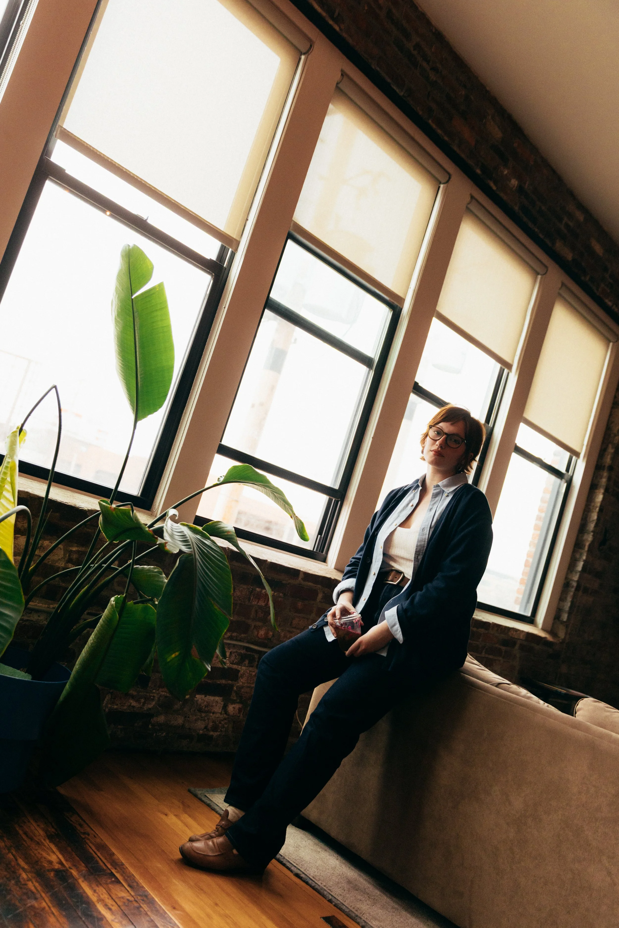 A woman with short brown hair, glasses, wearing a dark blazer and light-colored shirt, sitting on a beige sofa in a room with large windows and exposed brick wall, holding a glass of red beverage. There are large green houseplants near the window, an