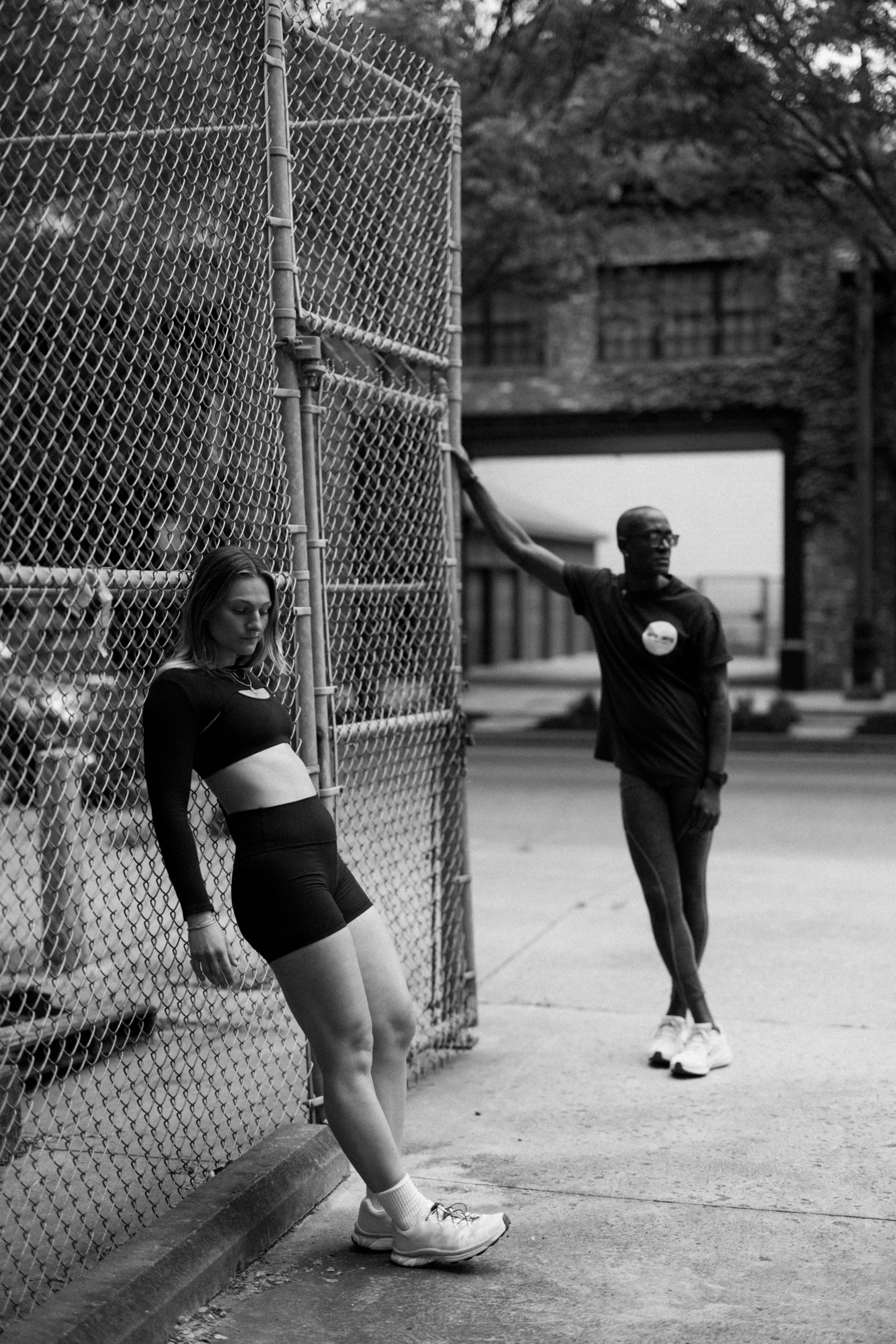 A young woman leaning against a chain-link fence, wearing athletic clothing and sneakers, with her head tilted downward. A man stands in the background, holding on to the fence with one arm, wearing glasses and casual athletic attire. They are outdoo