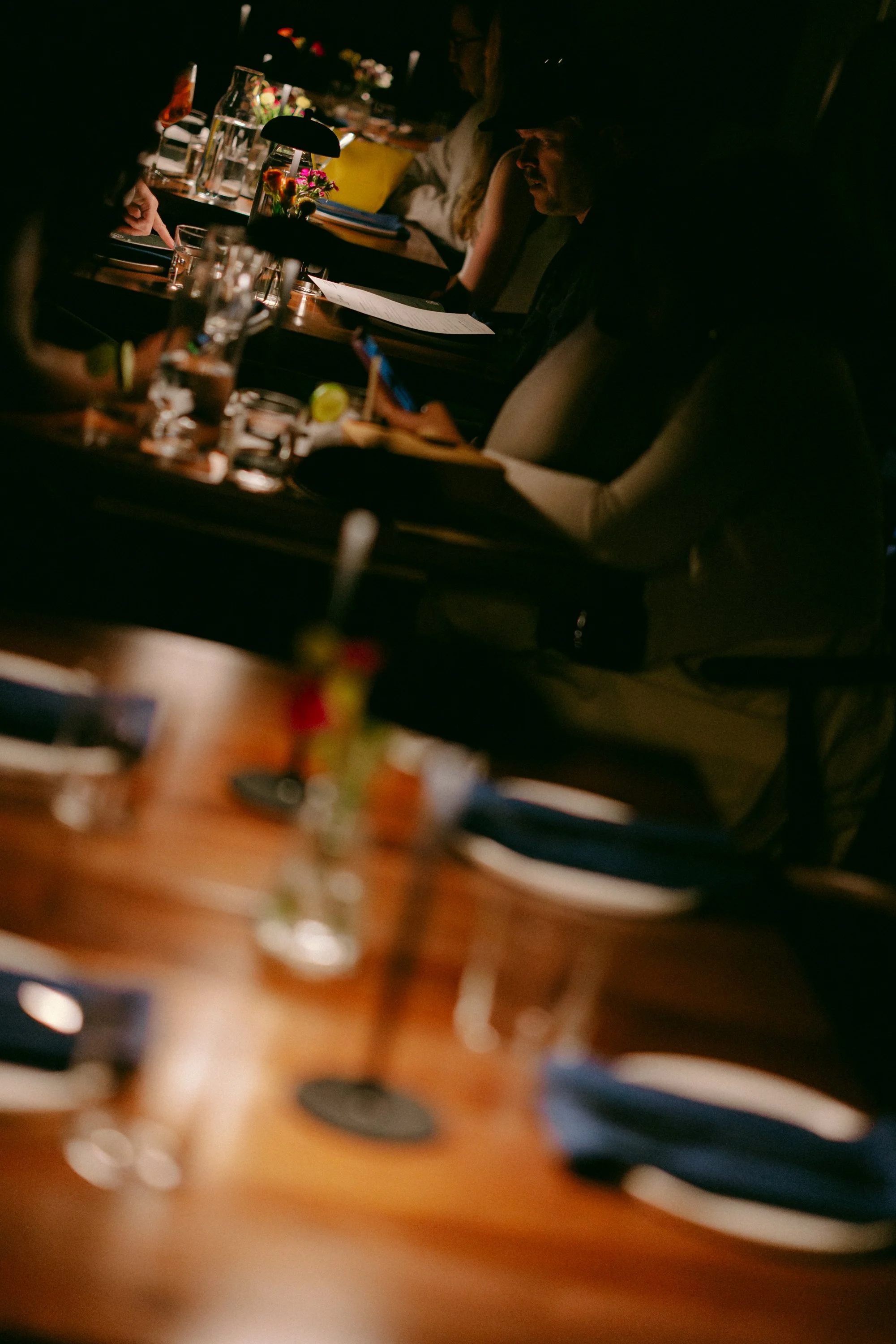 People sitting at a table in a dimly lit setting, with drinks, flowers, and papers in front of them, possibly at a casual meeting or social gathering.