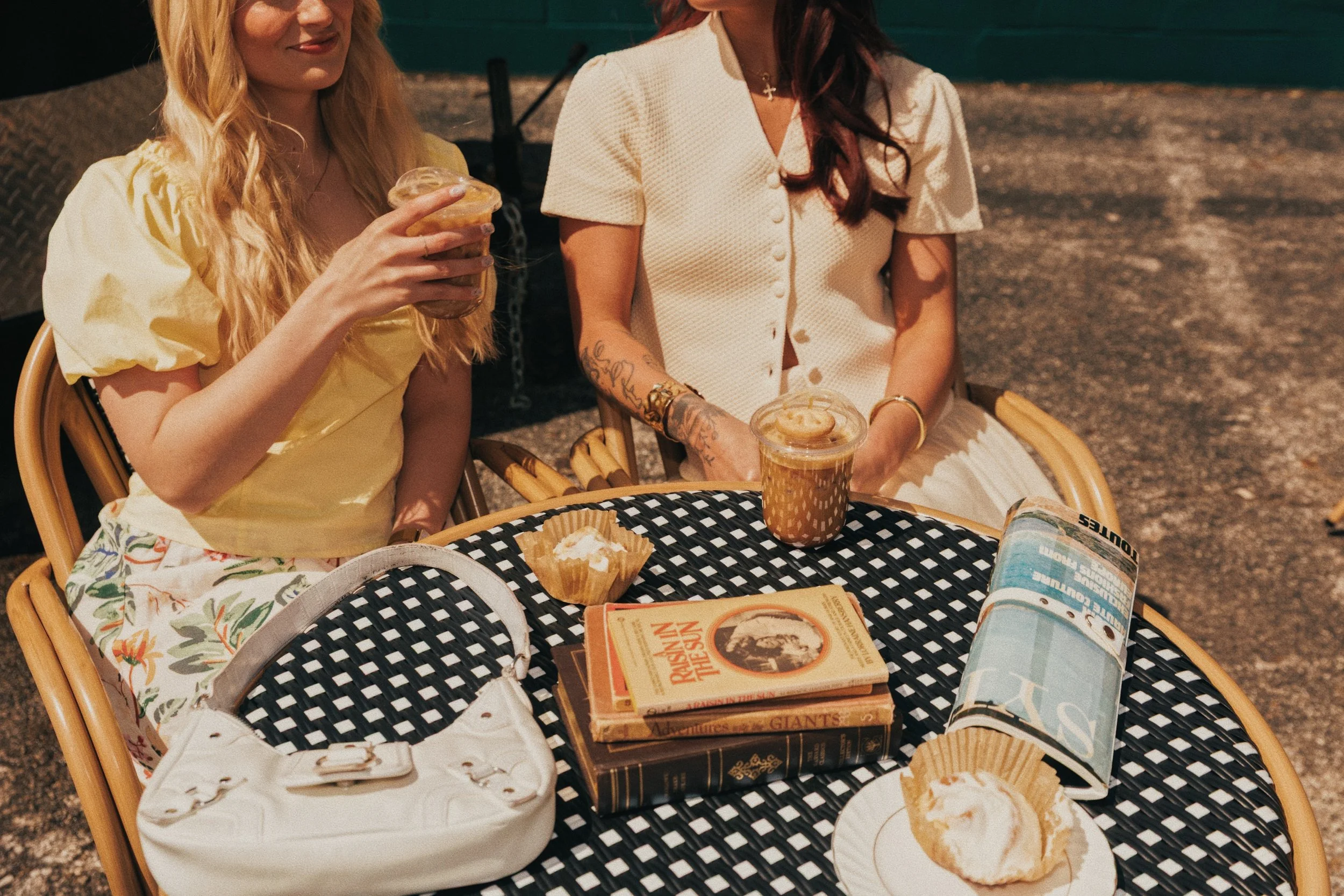 Two women seated at a round black and white checkered table outdoors, with drinks and snacks. One woman with blonde hair in a yellow dress is holding a caramel frappuccino, and the other with dark hair in a white dress has a similar drink. The table 