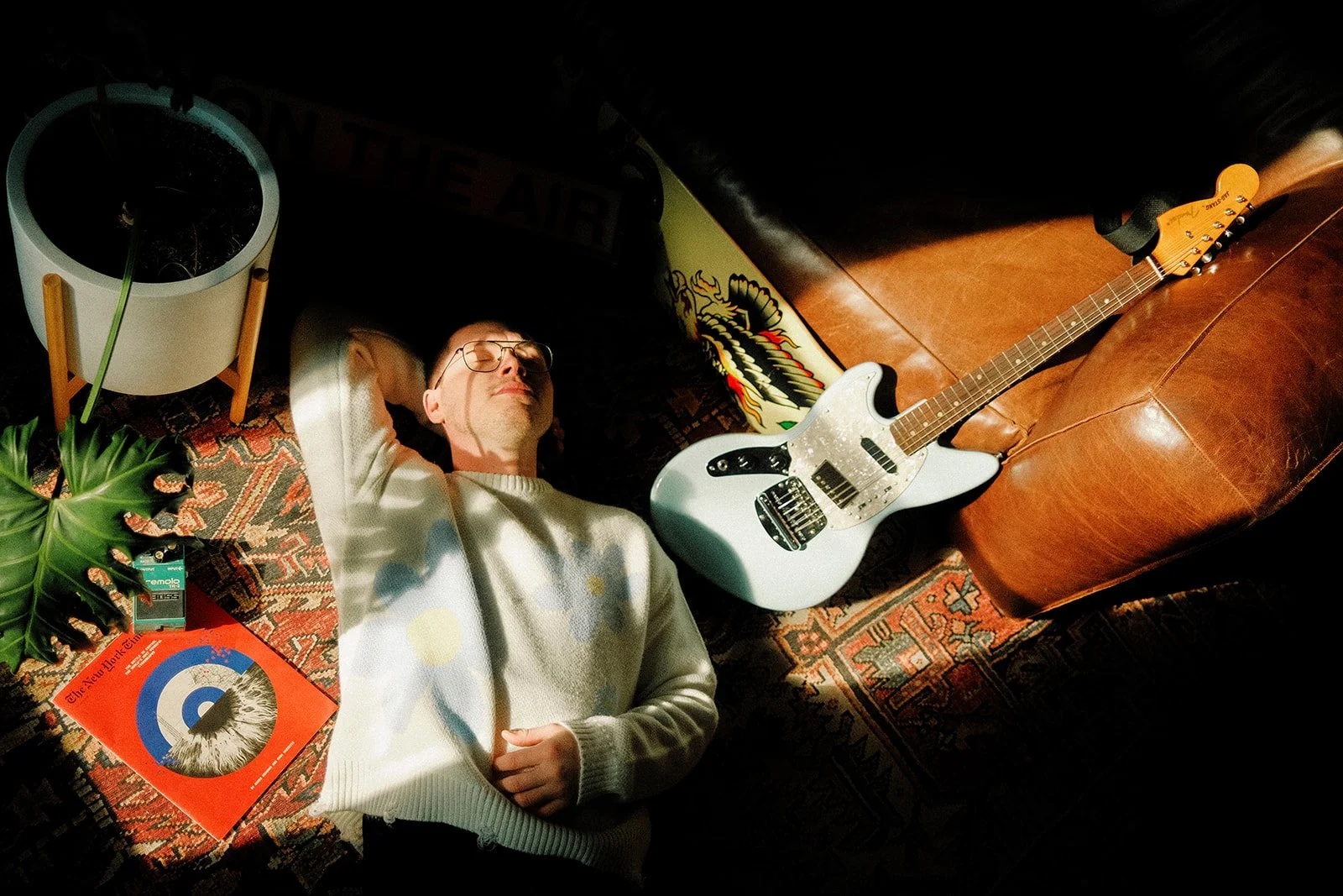 A man with glasses sleeping on a rug in a room, surrounded by a potted plant, a book, and an electric guitar, with sunlight casting shadows.