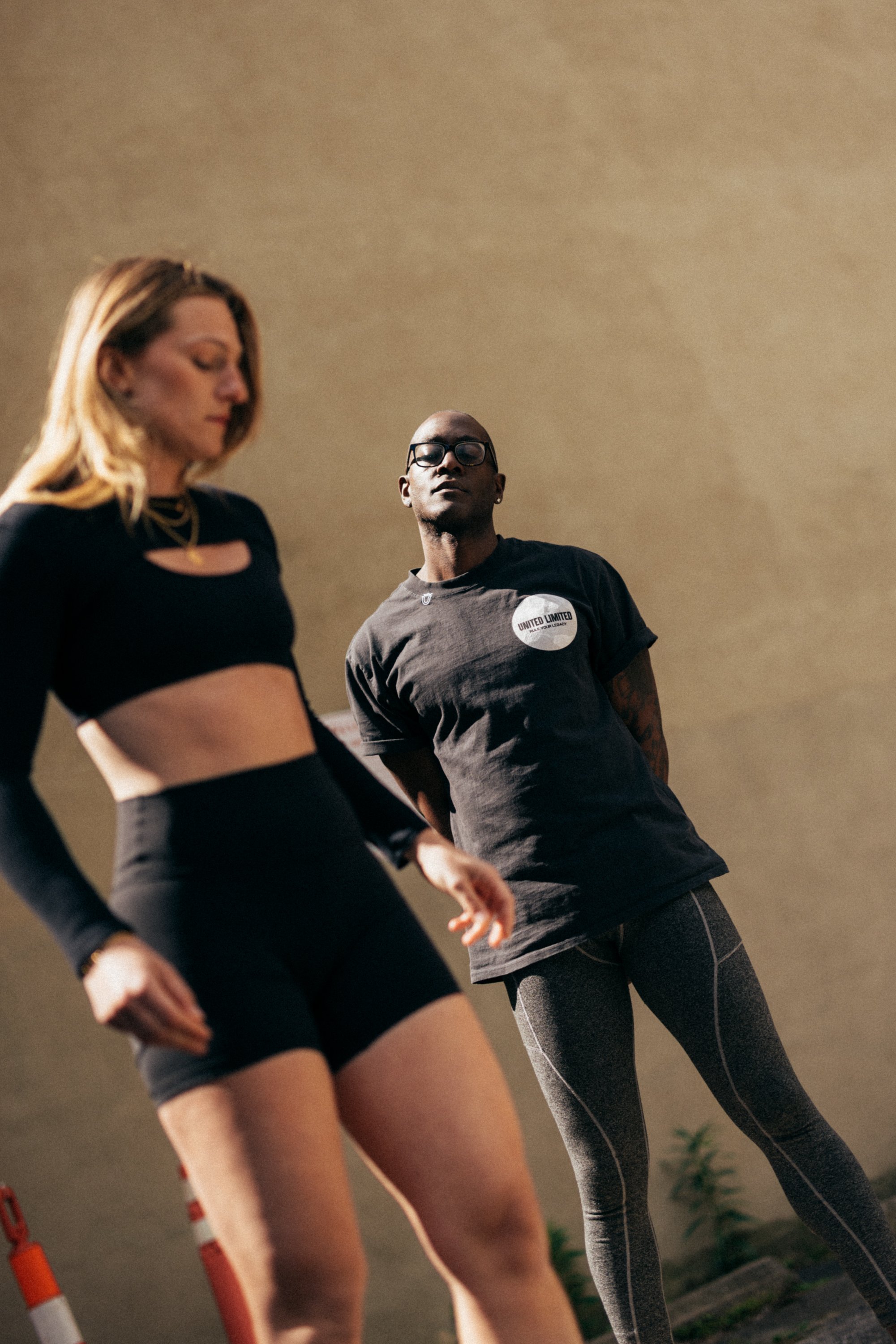 A woman with long blonde hair wearing a black sports top and shorts standing beside a man with shaved head, glasses, and tattoos wearing a dark gray T-shirt and gray leggings in a room with neutral walls and plants.