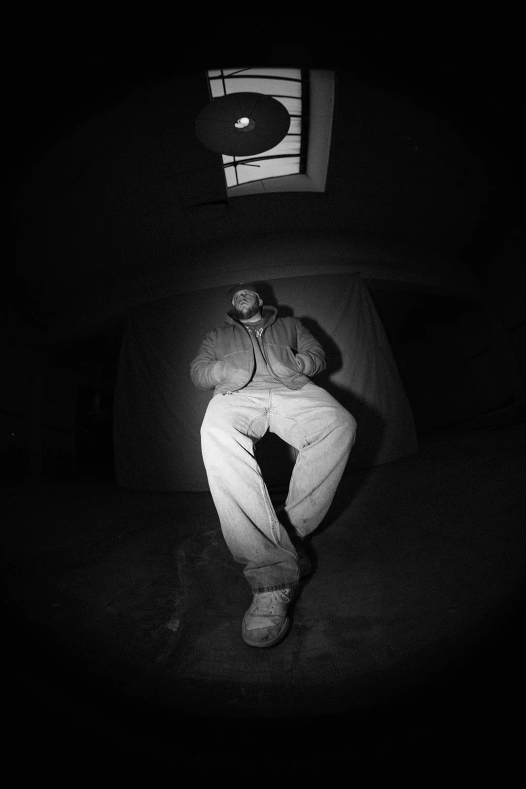 A man sitting with his hands in his jacket pockets, facing upward, wearing a jacket, hoodie, jeans, and sneakers, in a room with a wall behind him, a grid-patterned window, and a ceiling light fixture.