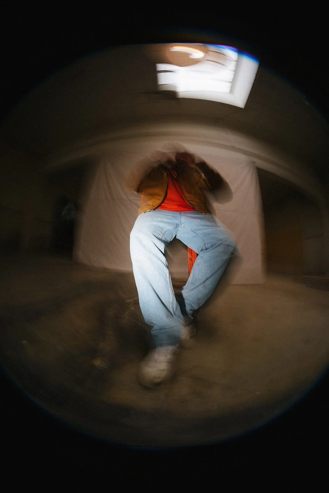 A person wearing a brown vest, red shirt, and blue jeans takes a selfie in a reflective surface with a fisheye effect, creating a distorted circular view.