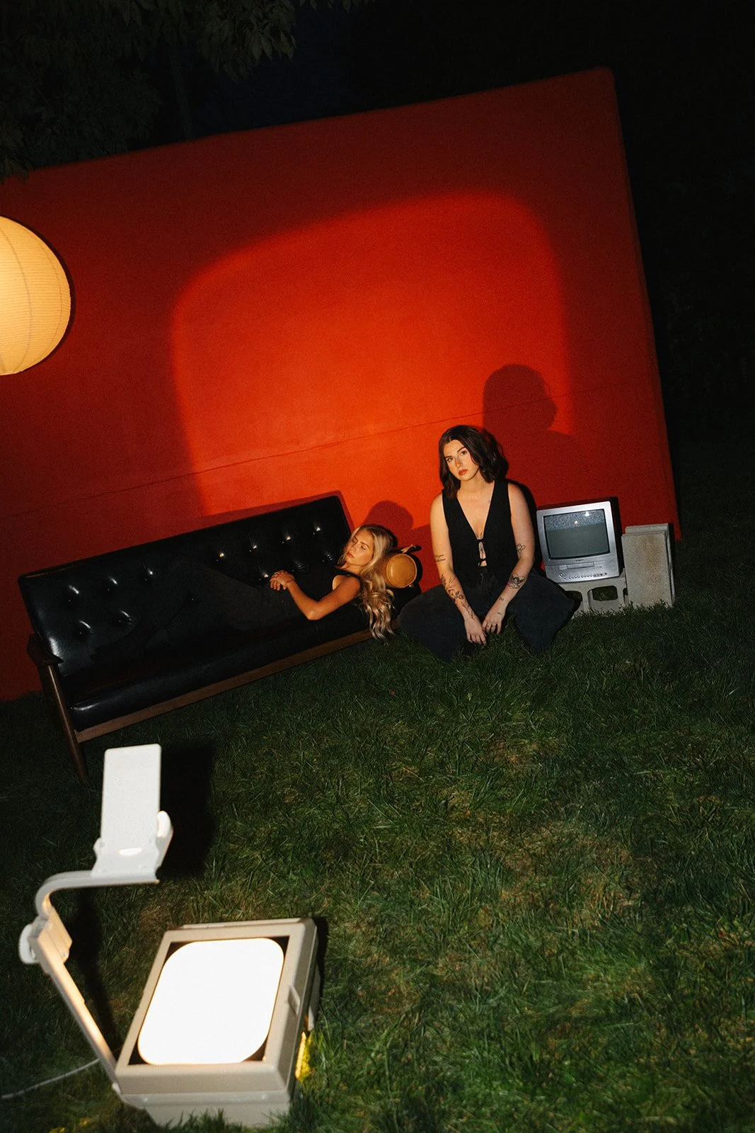 Two women in a dark outdoor scene, one lying on a black couch and the other sitting on grass, with a red wall, a vintage TV, a paper lantern, and studio light.