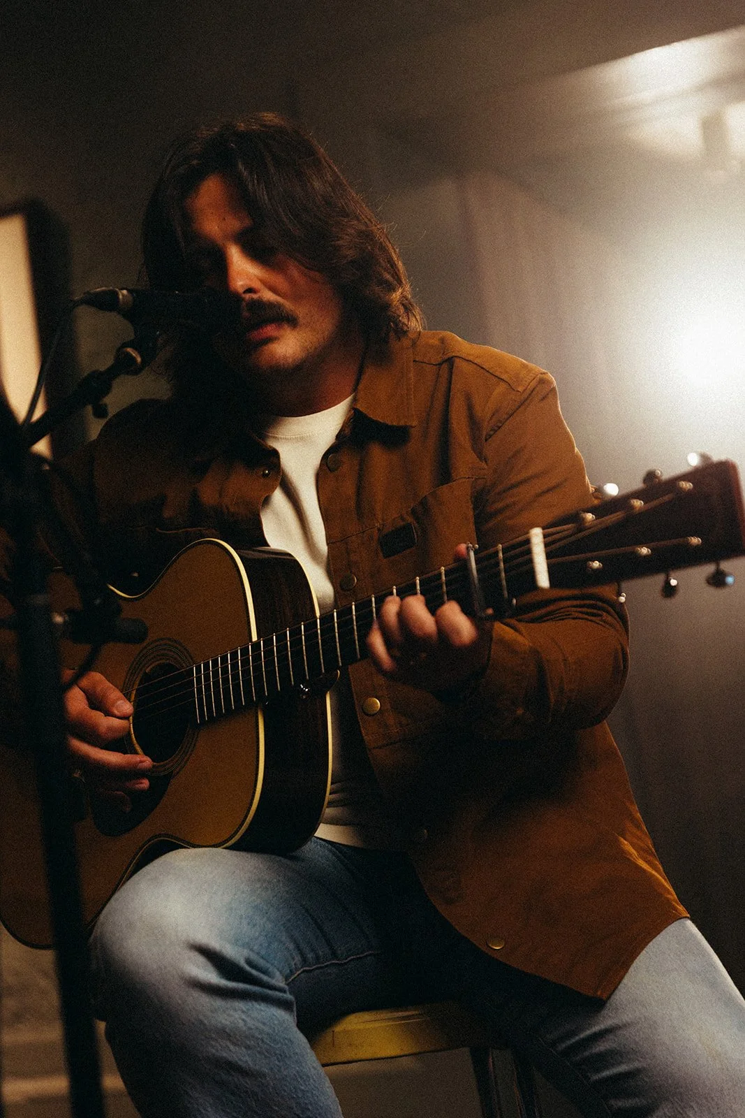 A man with long hair and a mustache playing an acoustic guitar and singing into a microphone in a dimly lit room.