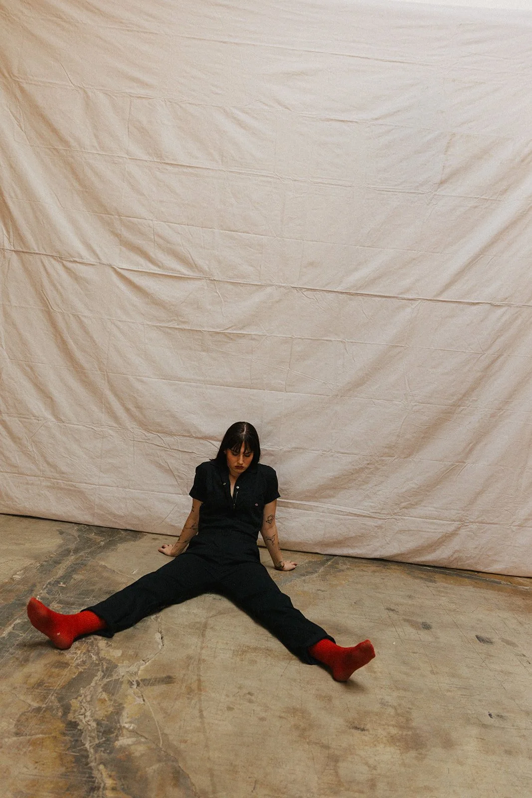 A woman with black hair and tattoos sitting on the floor with legs spread wide apart, wearing a black outfit and red socks, against a beige backdrop in an indoor setting.