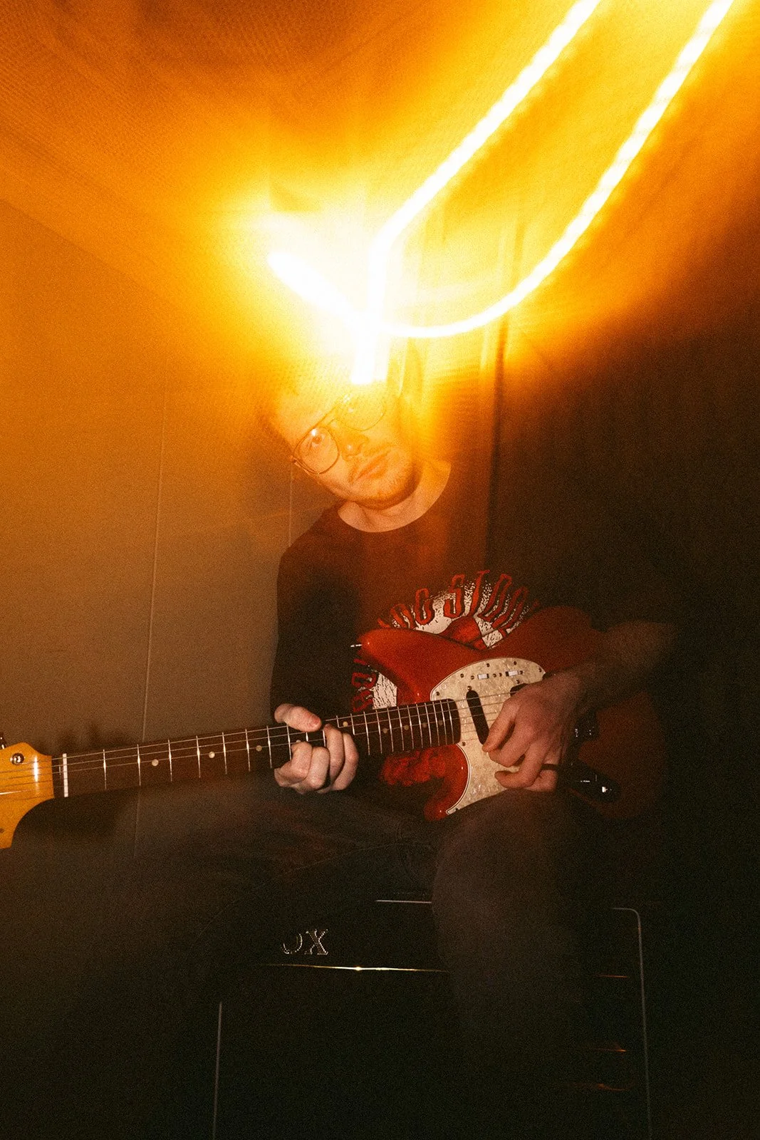 A man wearing glasses and a black t-shirt with a red and white graphic playing an electric guitar in a room with dim lighting and a bright light source above him.