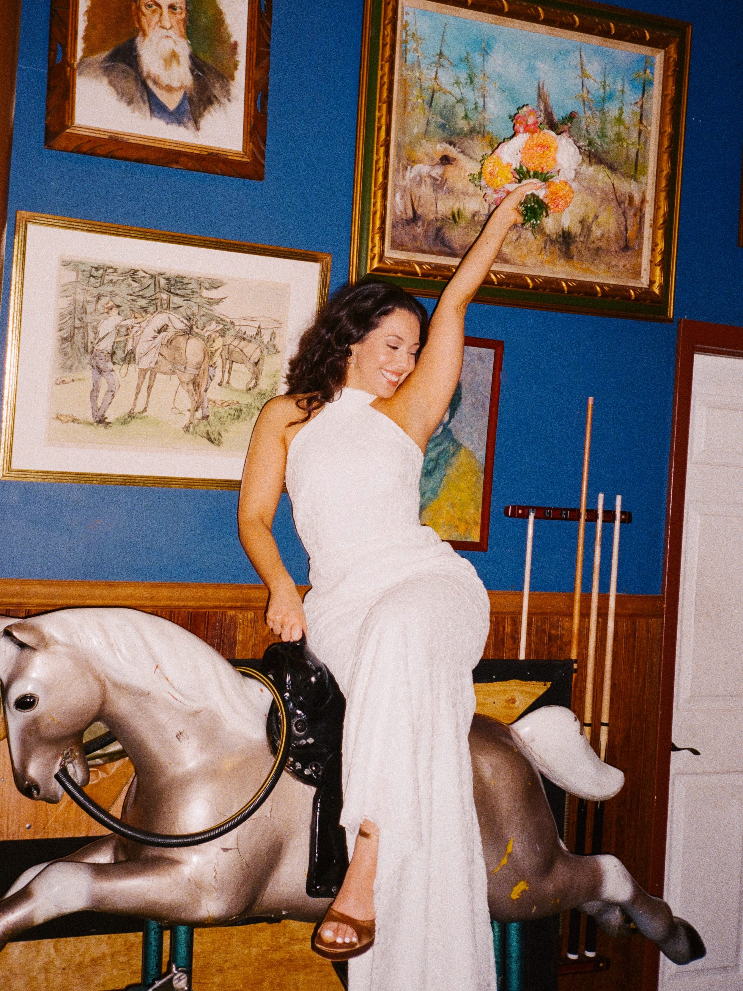 A woman in a white dress is sitting on a mechanical horse in an art gallery. She is smiling and holding a bouquet of flowers in her raised right hand. The gallery walls are blue and decorated with various paintings, including a portrait of a bearded 