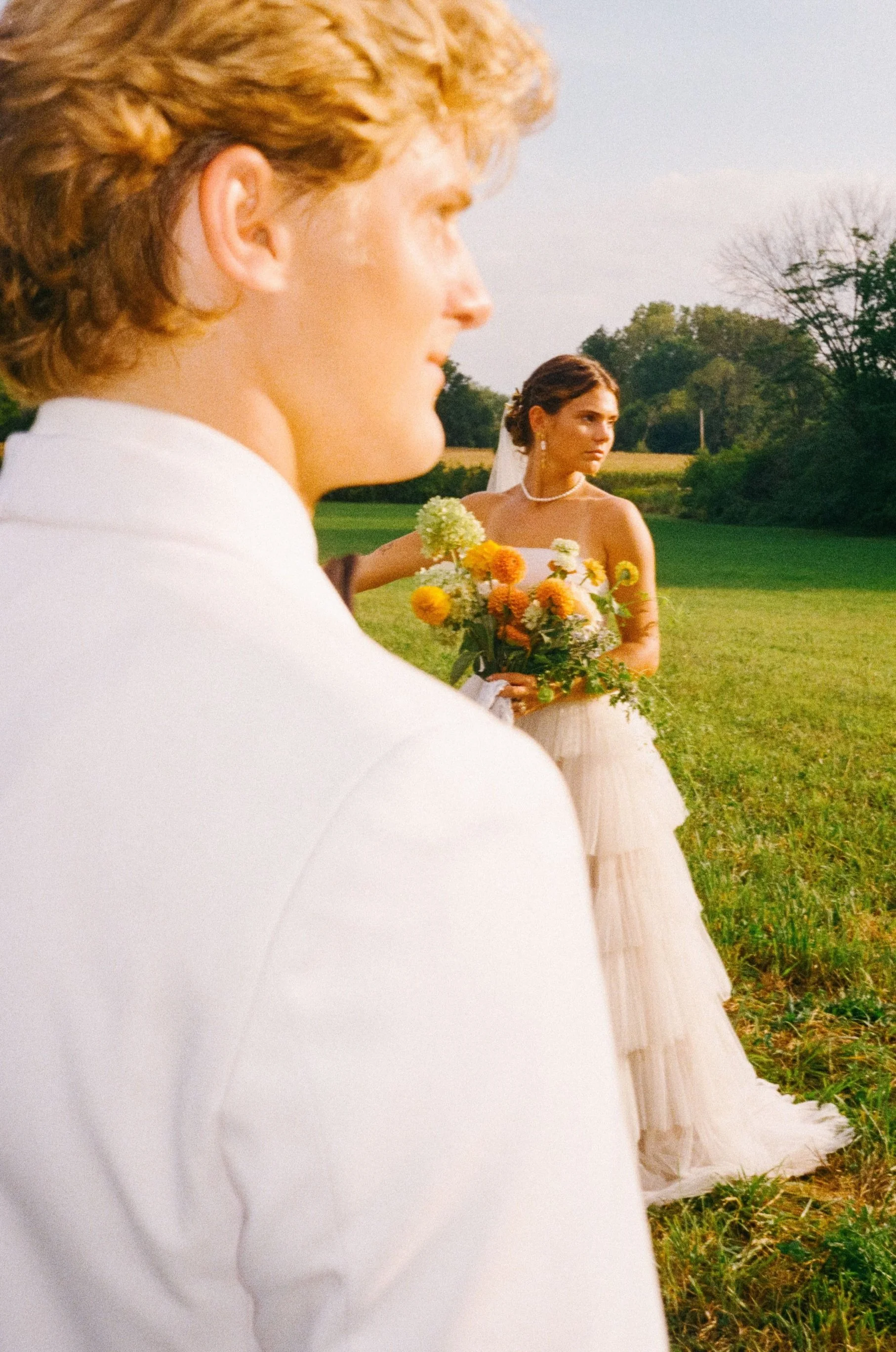 A man and woman dressed in wedding attire outdoors in a field, with the man in the foreground and the woman in the background holding a bouquet of yellow, white, and orange flowers.