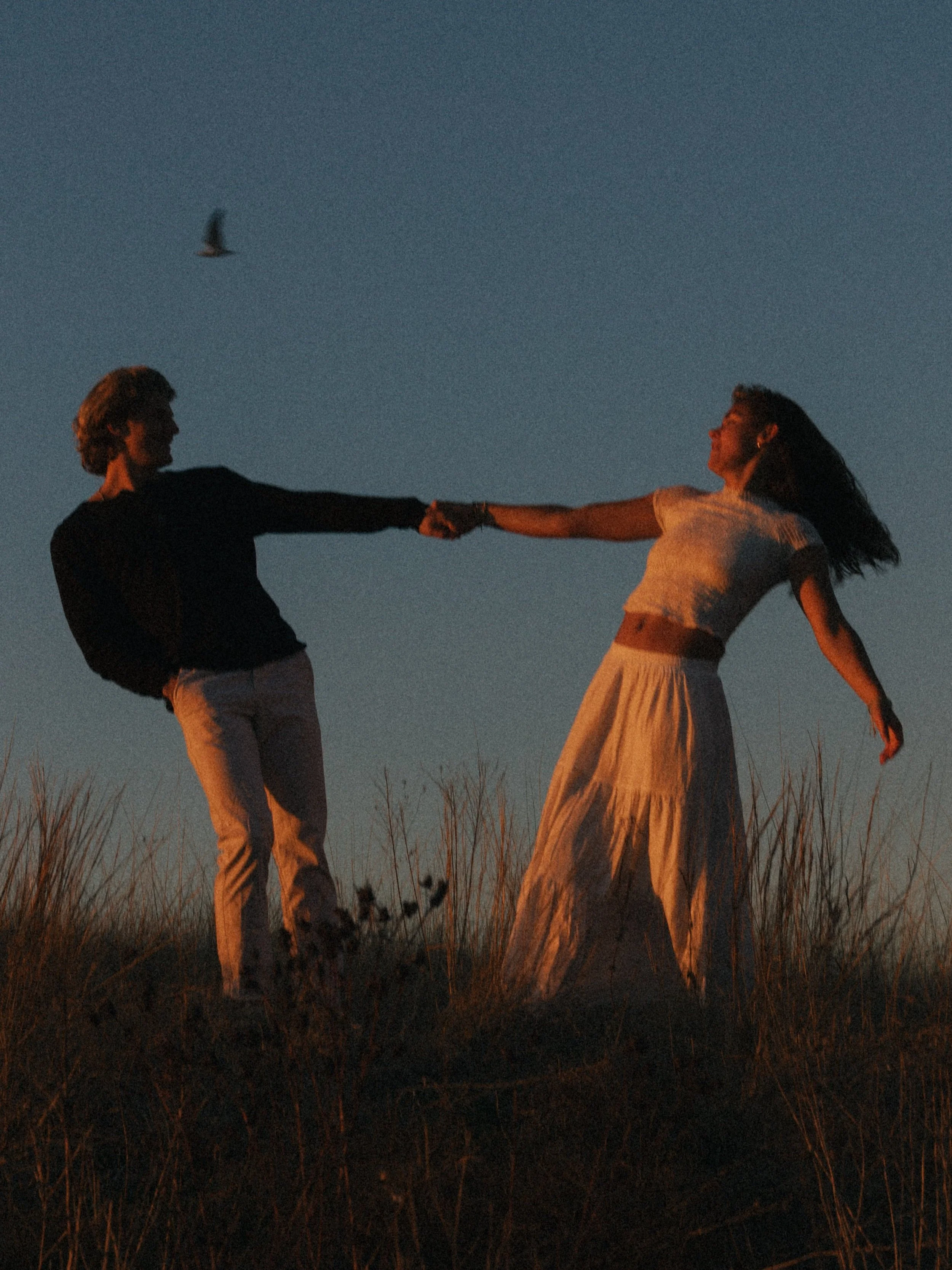 A couple holding hands and dancing on a grassy hill at sunset, with a bird flying in the sky.