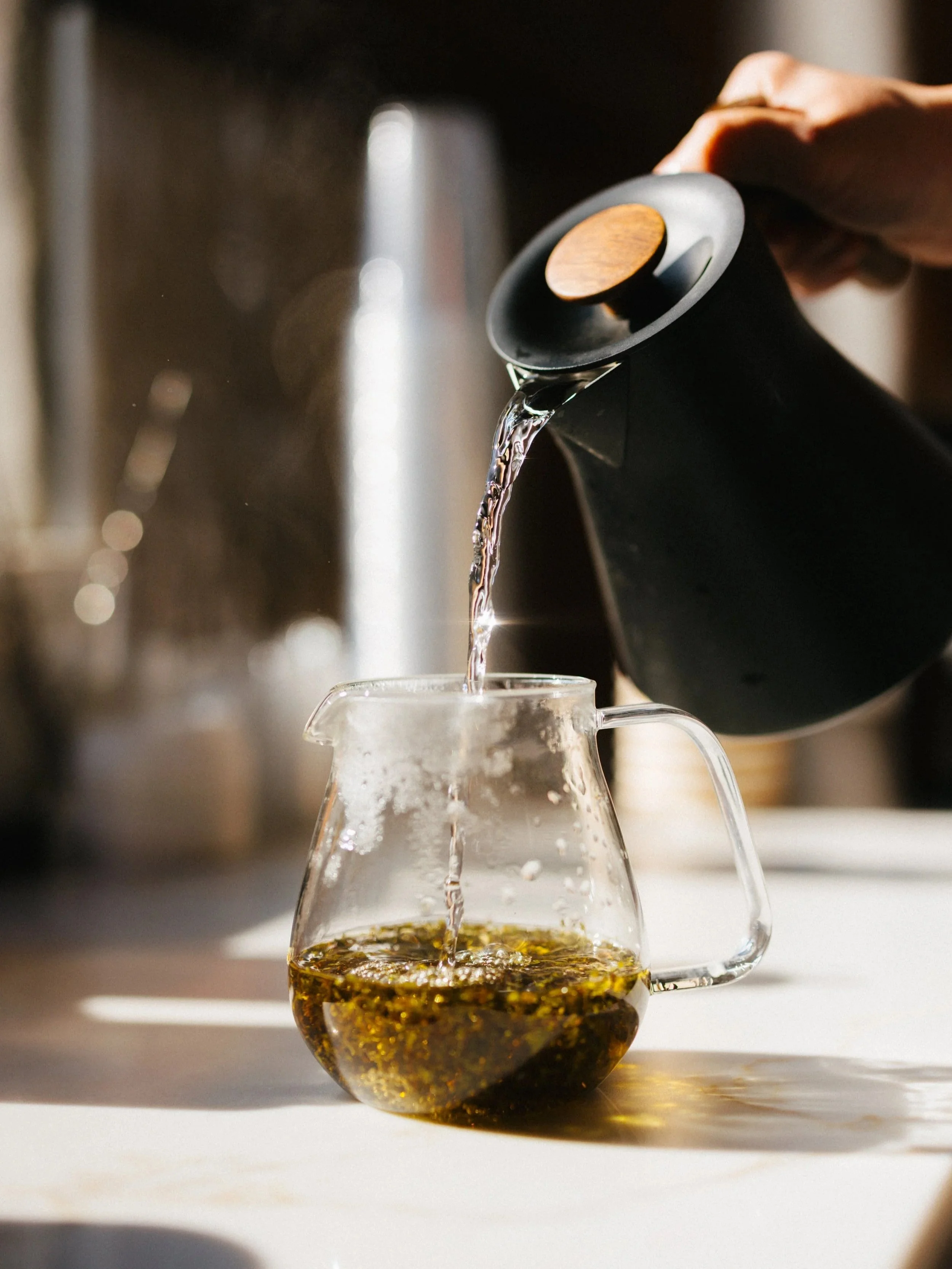 Pouring hot tea from a black teapot with wooden handle into a glass teapot containing loose herbs or tea leaves.