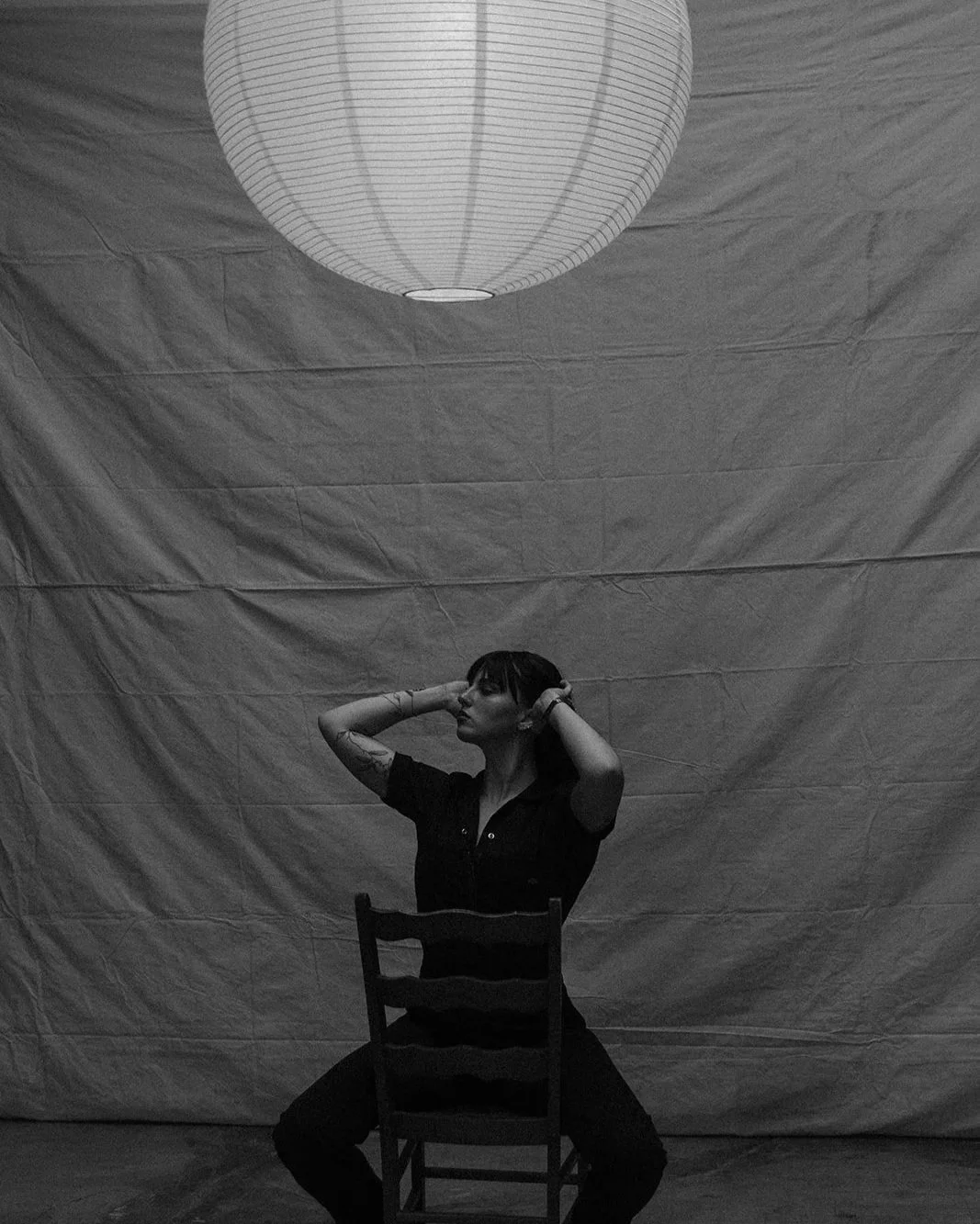 A woman sitting on a chair facing forward, with a large paper lantern hanging from the ceiling above her. The background is plain and textured. The woman has short hair and tattoos, and she is holding her hands behind her head.